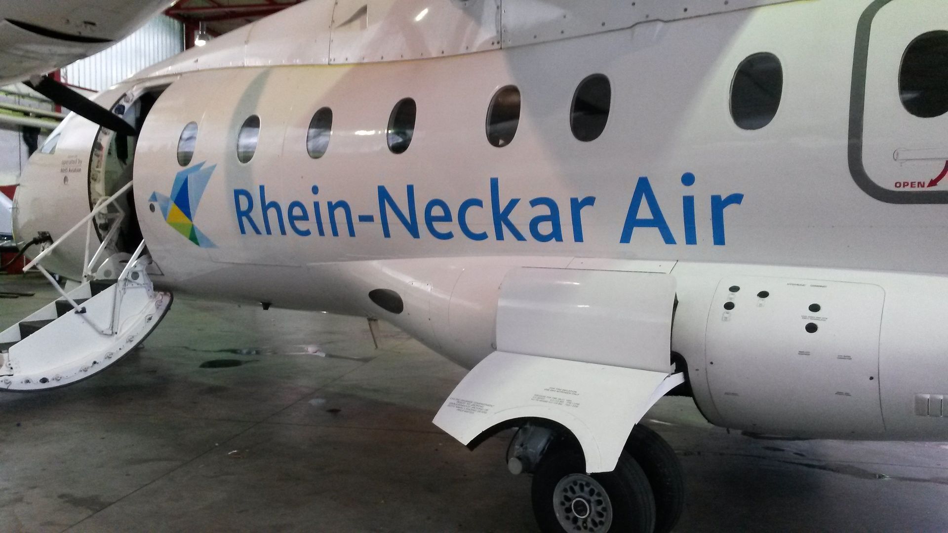 Seite eines weißen Flugzeugs der Rhein-Neckar Air mit blauer Beschriftung und offener Tür in einem Hangar.