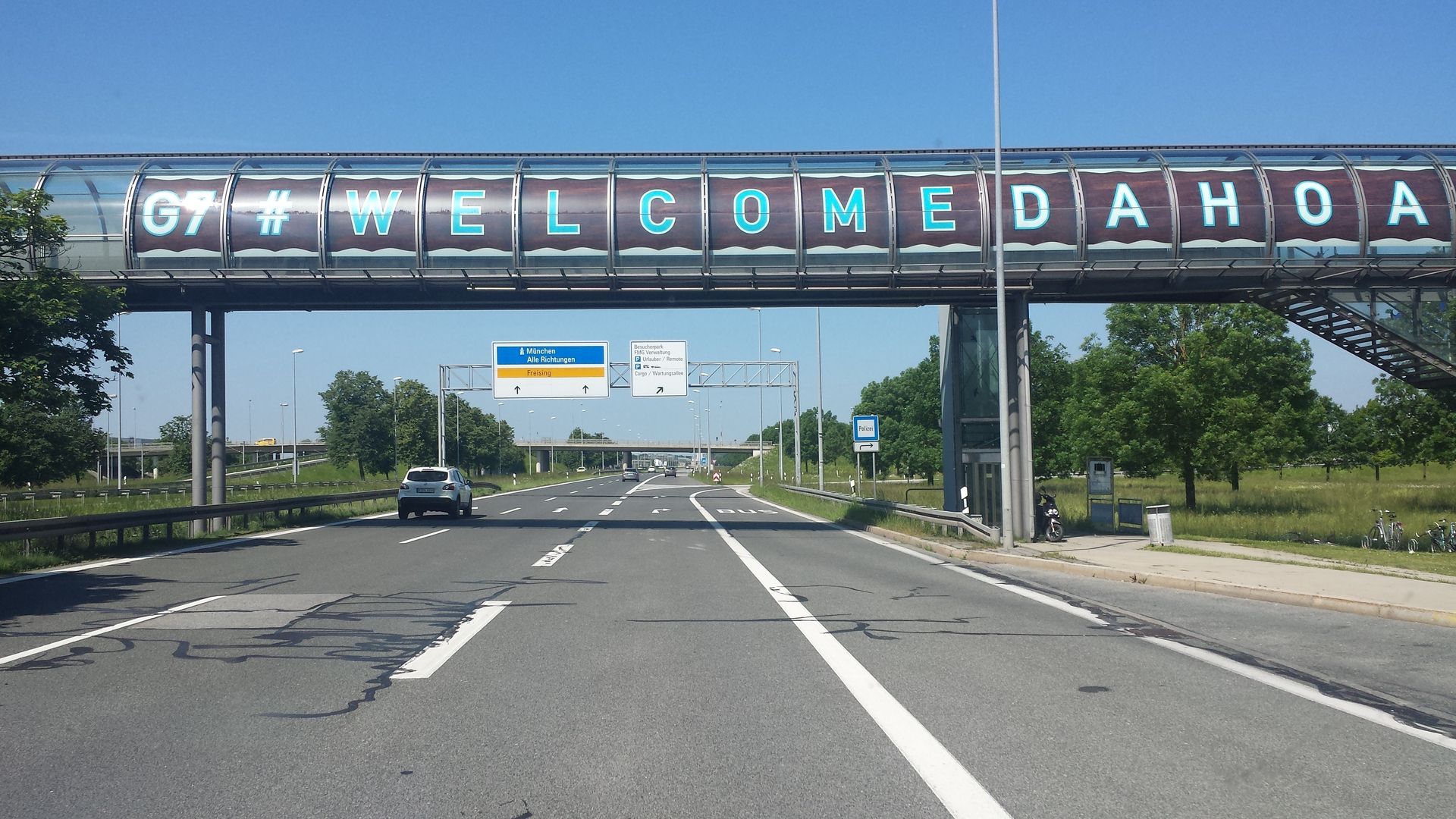 Autobahn unter einer Brücke mit dem Schild „WELCOME DAHOA“; ein Auto fährt unter einem sonnigen Himmel auf der Straße.