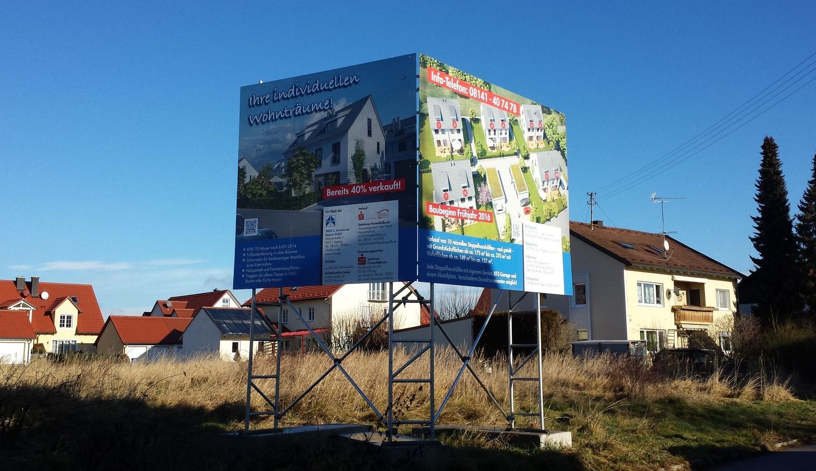 Plakatwand mit Werbung für neue Häuser; auf einem Stahlgerüst auf einem Feld platziert, im Hintergrund Häuser.