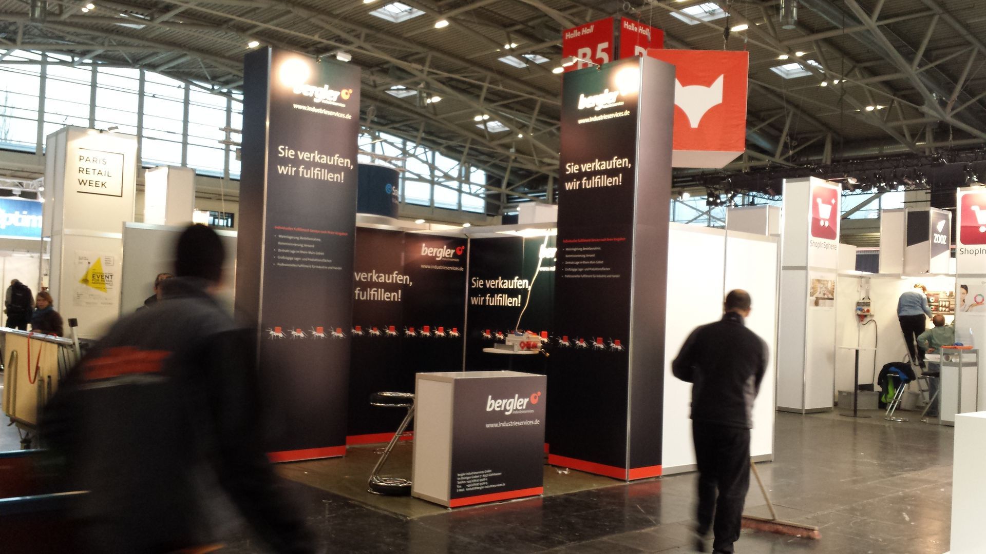 Messestand mit dunkelrotem und schwarzem Branding. Menschen gehen vorbei.