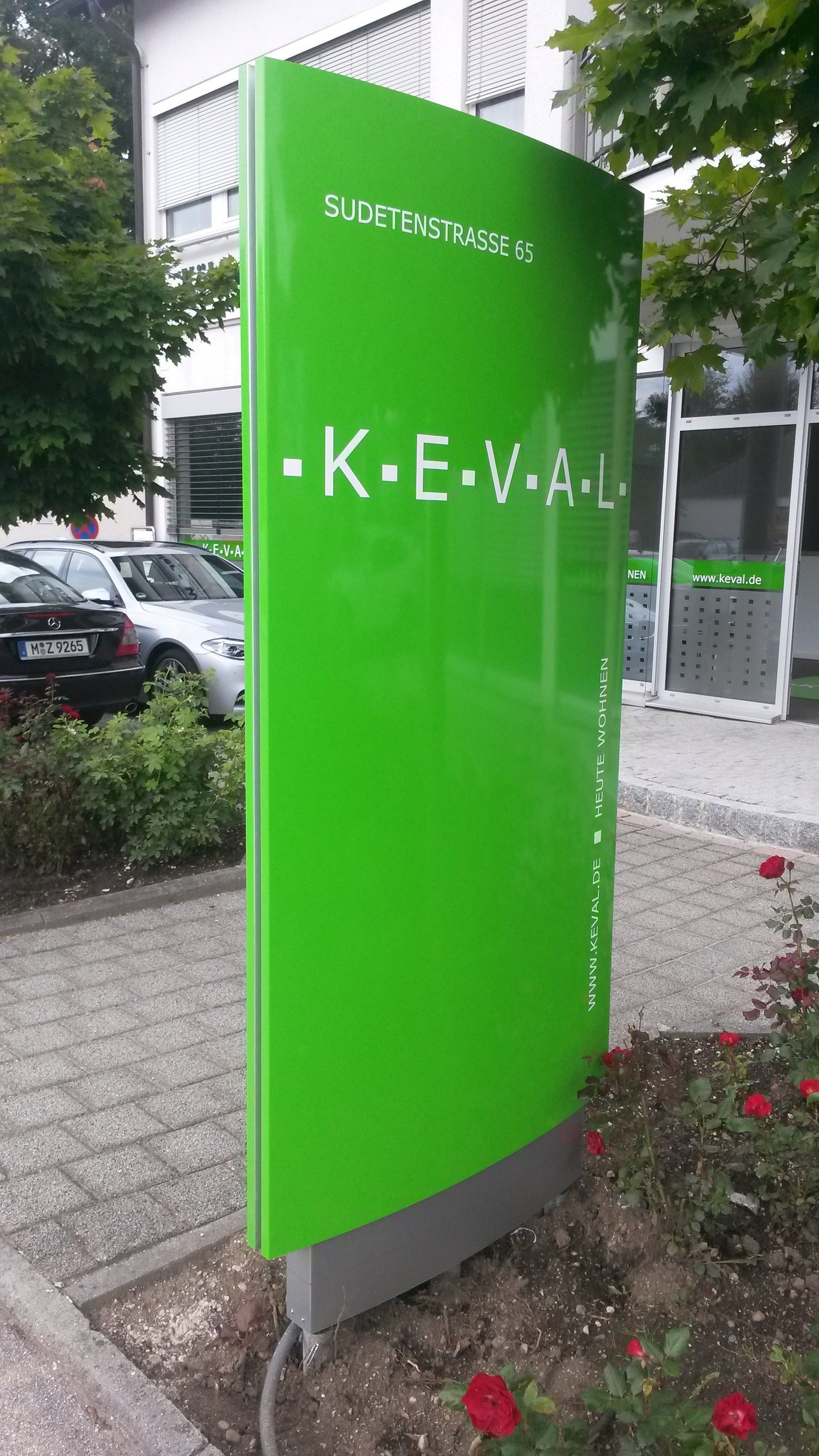 Grünes Schild der Firma „K•E•V•A•L“ vor dem Gebäude, Autos im Hintergrund.