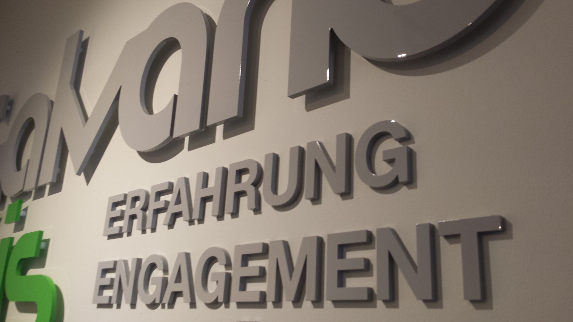 Graue 3D-Buchstaben an einer Wand, die „ERFAHRUNG ENGAGEMENT“ unter einem teilweise sichtbaren Logo buchstabieren.