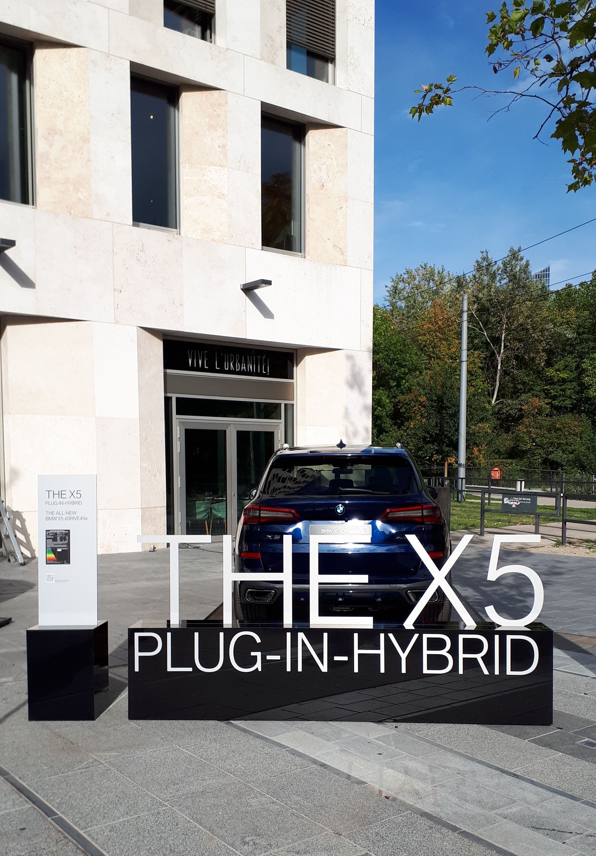 Blauer BMW X5 Plug-in-Hybrid, geparkt vor einem Gebäude mit weißem Schild.