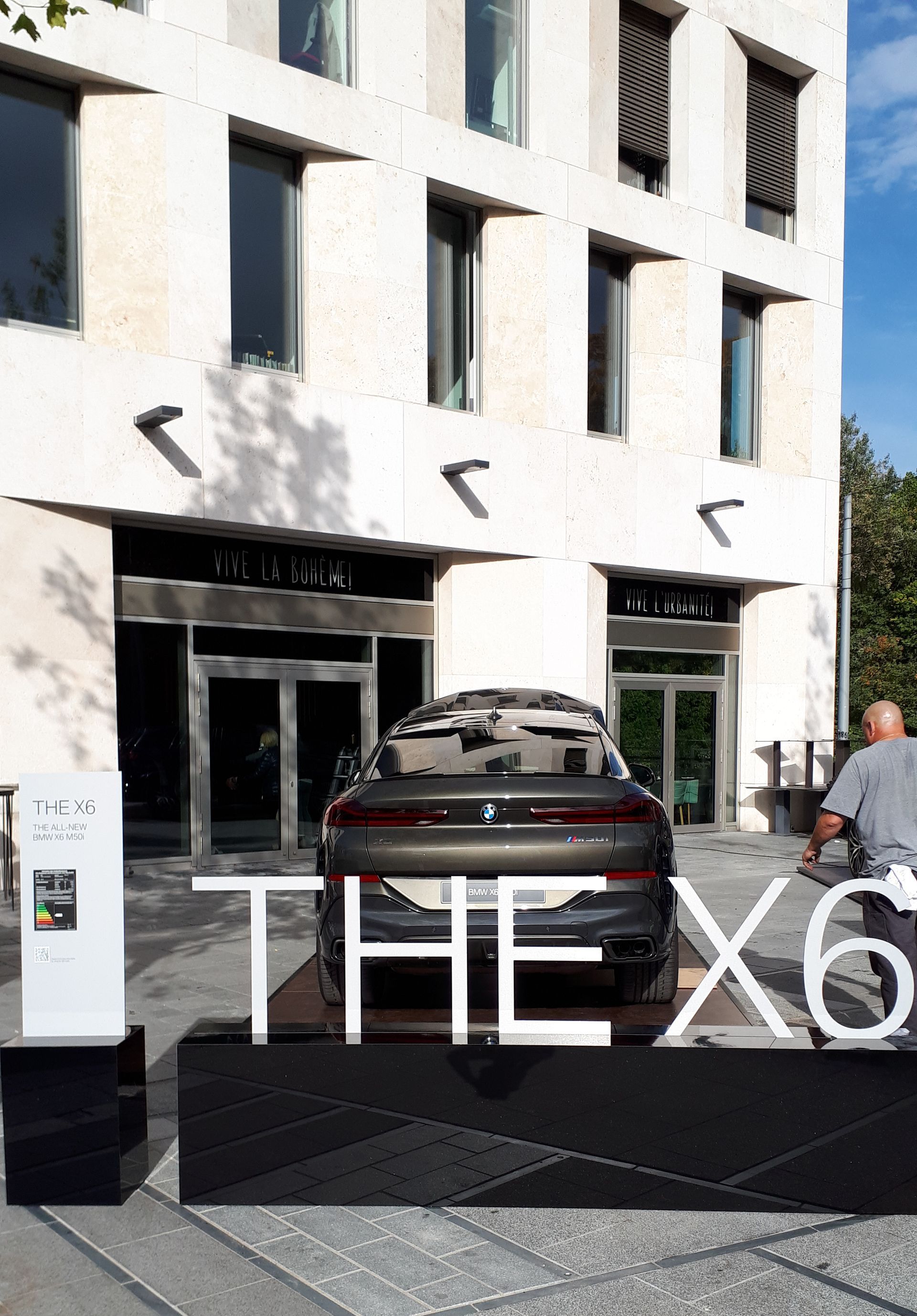 Vor einem Gebäude steht ein BMW X6 mit einem großen Schild mit der Aufschrift „THE X6“. Ein Mann geht rechts.