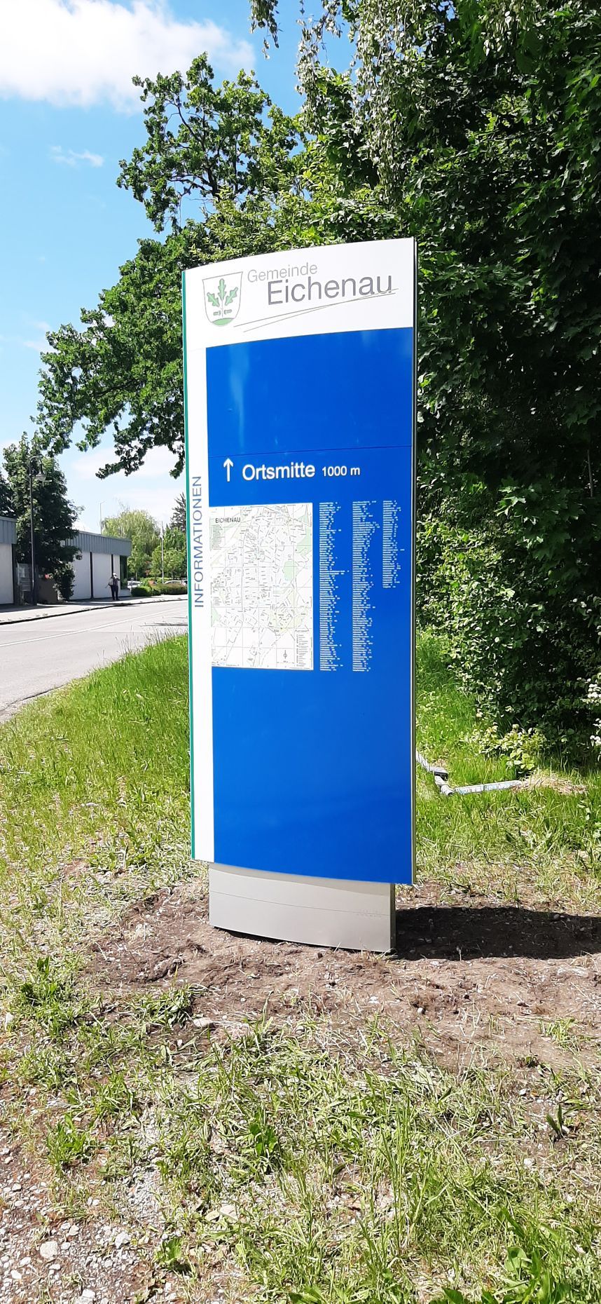 Wegweiser auf einer Rasenfläche, blau-weiß, mit dem Ortsnamen Eichstetten. Im Hintergrund Bäume und Himmel.