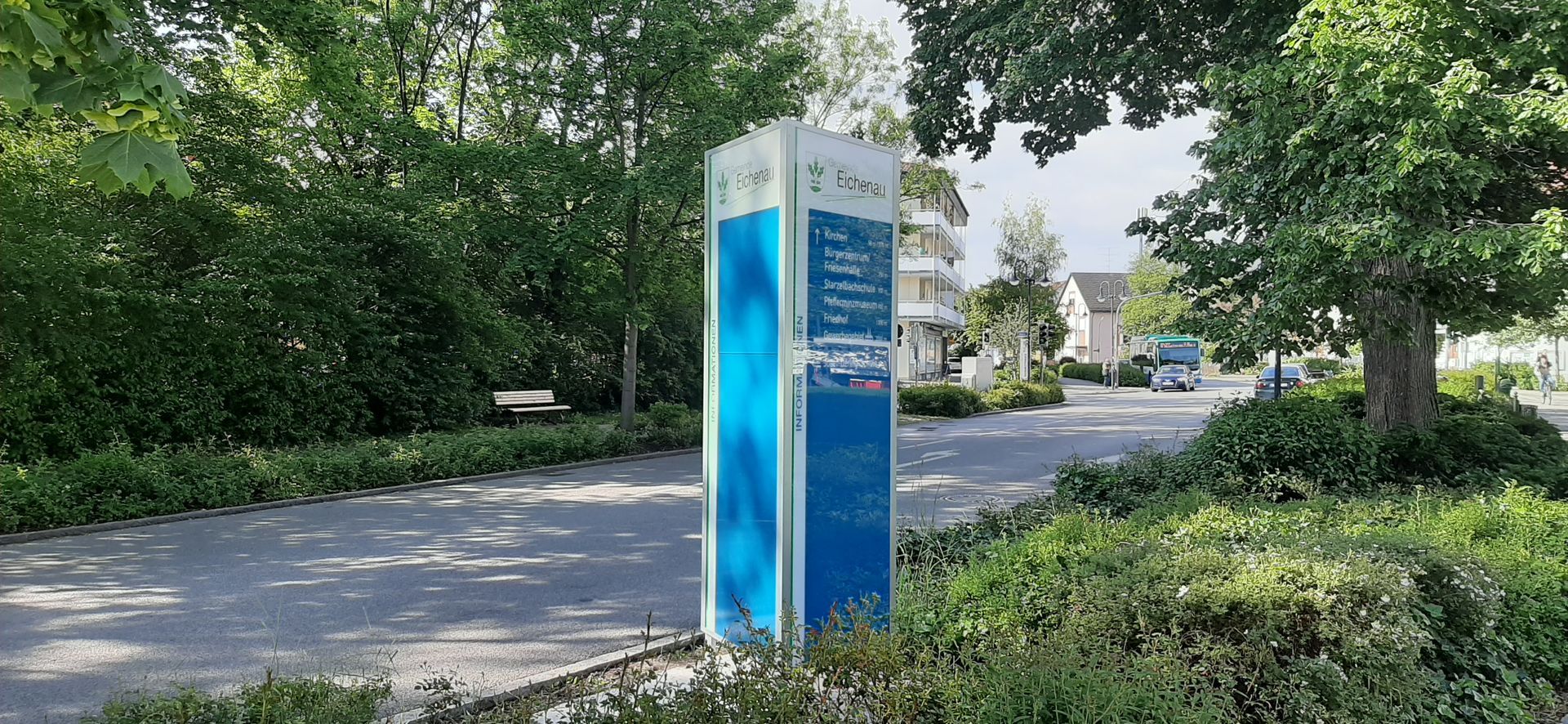 Ein blauer Werbepylon steht an einer Straße neben Büschen und Bäumen.