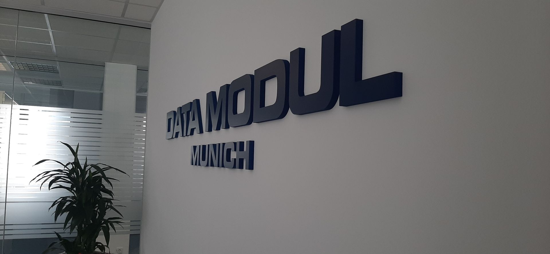 DATA MODUL-Beschilderung an einer weißen Wand mit einer Pflanze und einem Glasfenster im Hintergrund.