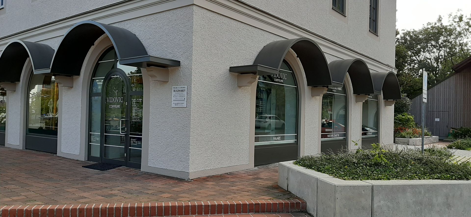 Gebäude mit Bogenfenstern und Markisen; davor ein Bürgersteig aus Ziegeln und ein niedriger Blumenkasten aus Beton.