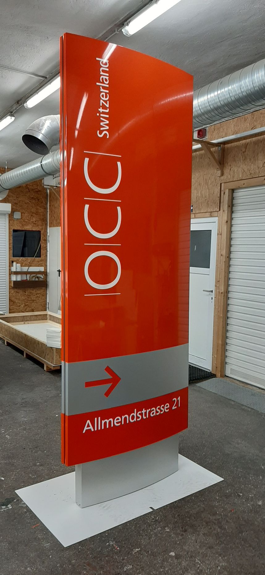 Orange-graues Wegweiserschild in einer Werkstatt. Zeigt einen Pfeil und den Text „OCCC, Almendsási 21.“
