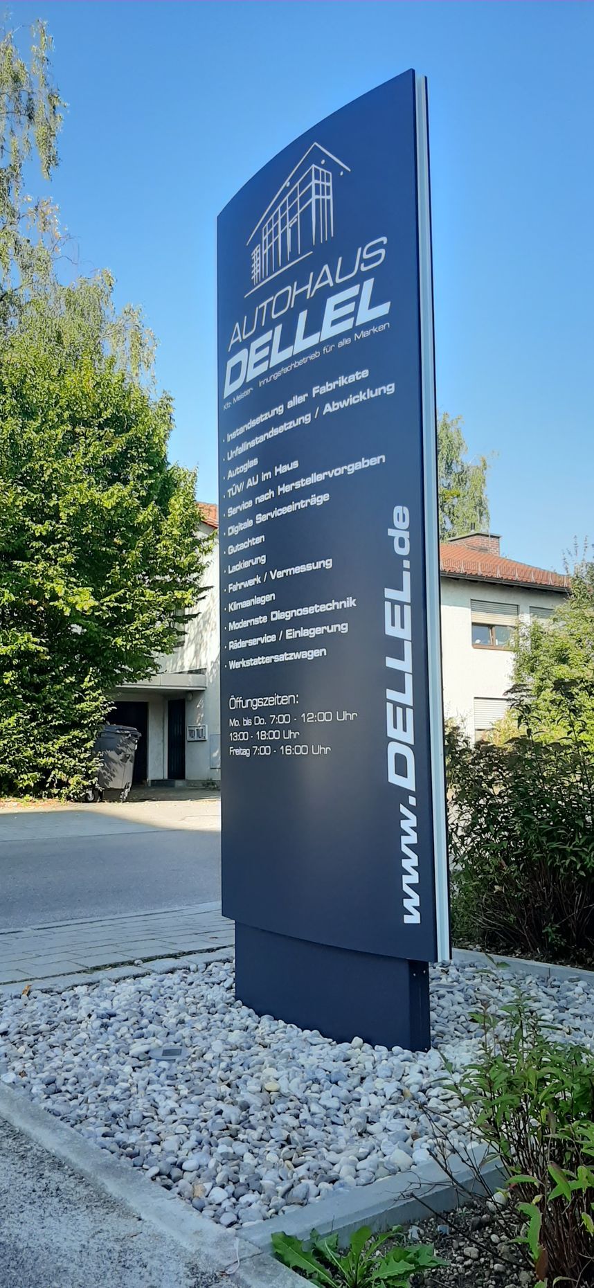 Ein hohes, dunkles Schild für Autohaus Dellel mit Text und Logo, aufgestellt vor einem Gebäude und Kies.
