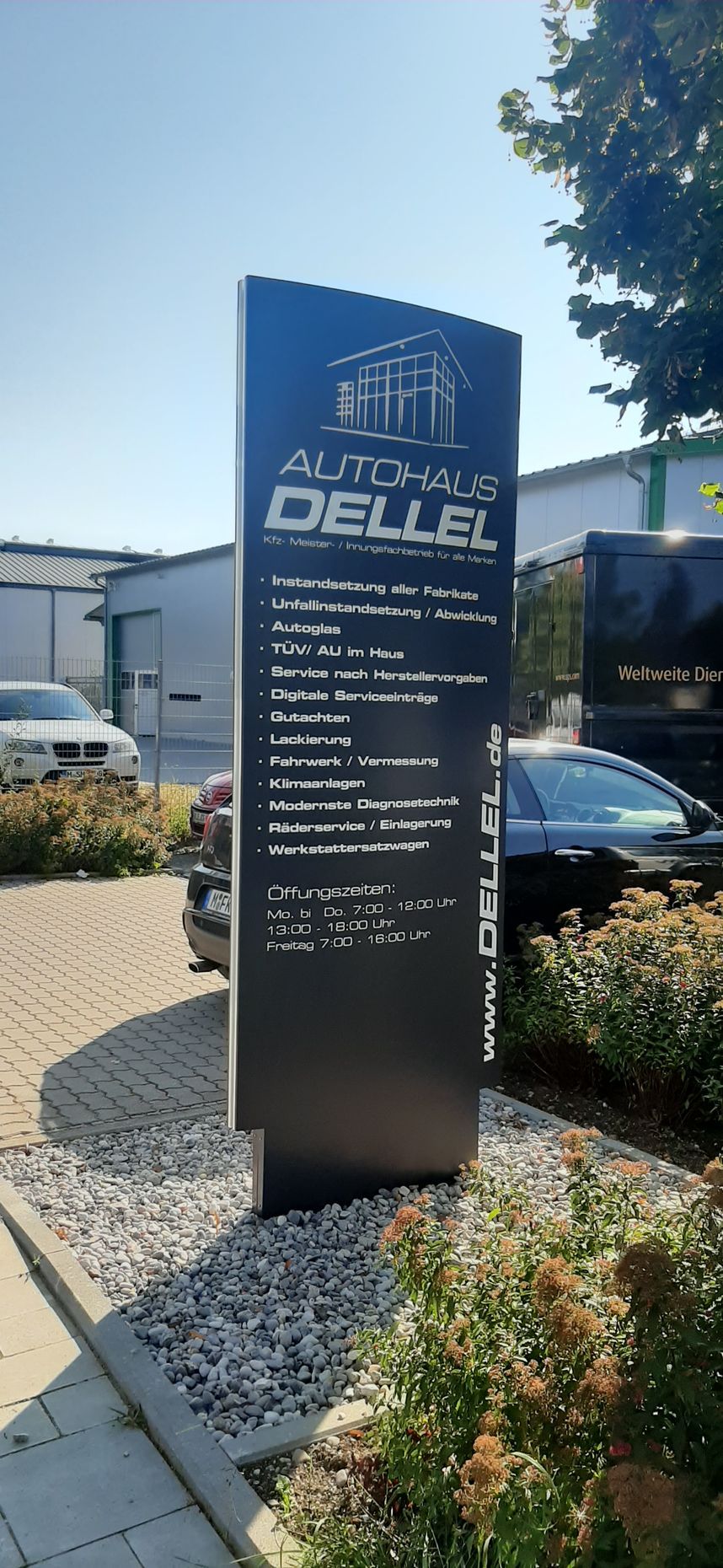 Neben einem Gebäude steht ein schwarzes Firmenschild mit der Aufschrift „Autohaus Dellel“, der Himmel ist blau.