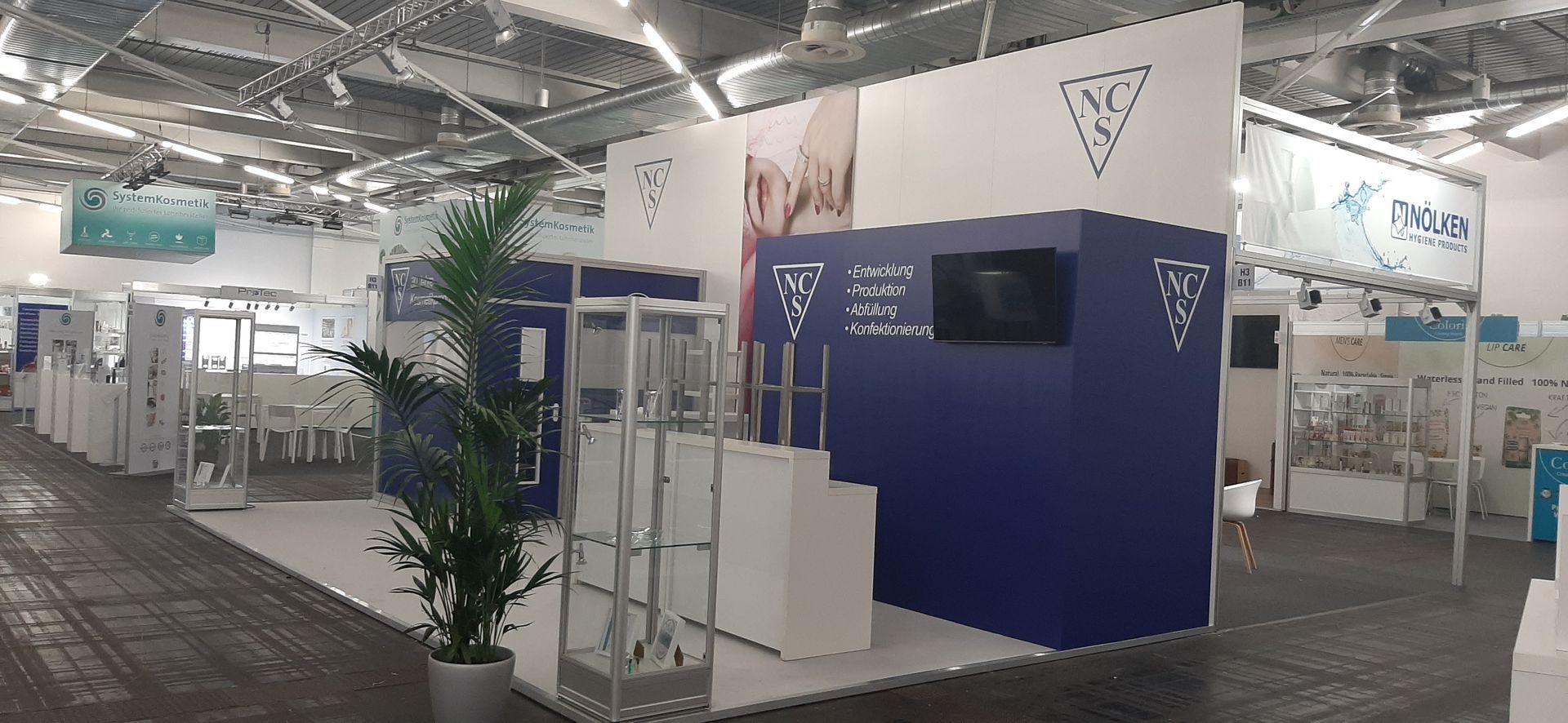 Eine Ausstellungshalle mit mehreren Ausstellungsständen. Im Mittelpunkt steht ein blau-weißer Stand mit Logo.