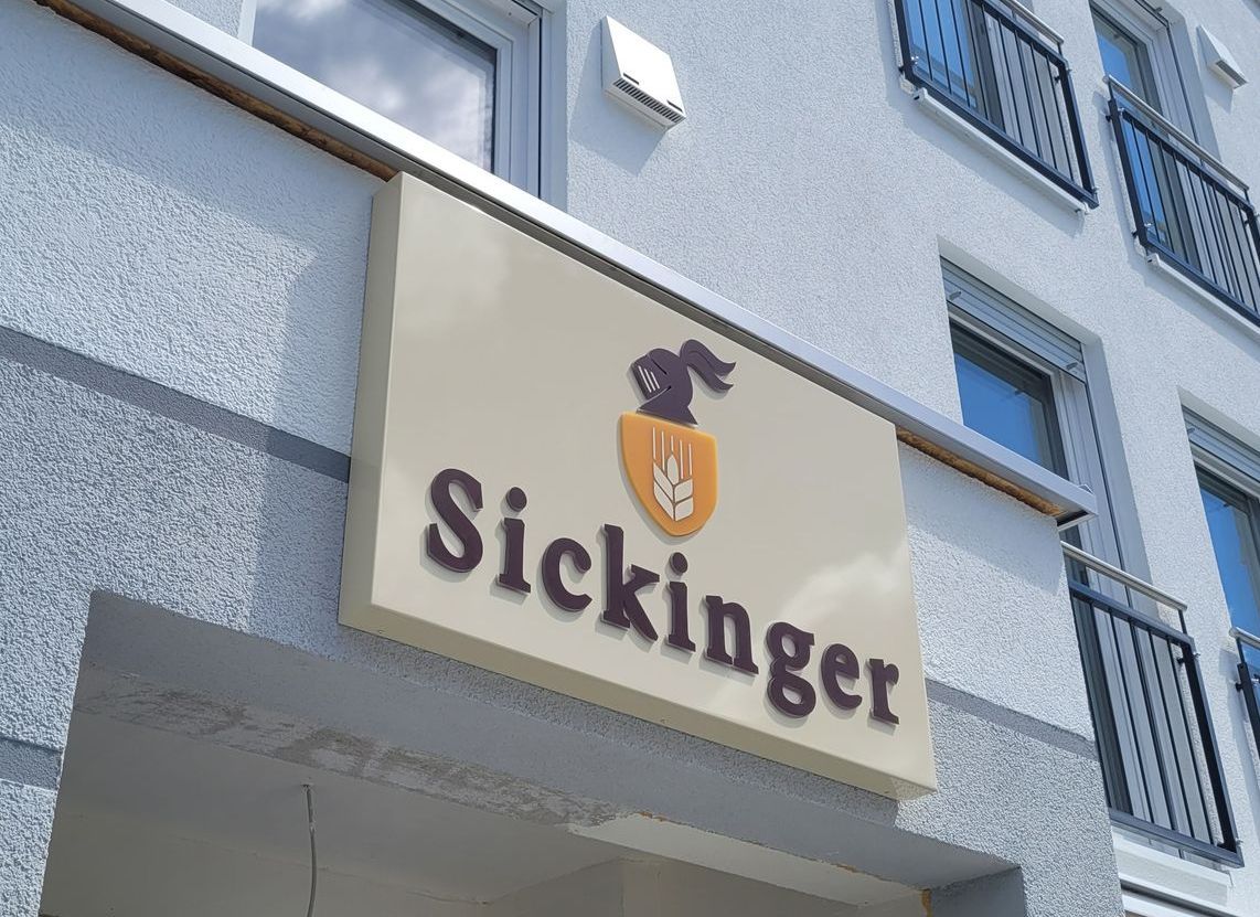 Leuchtkasten der Firma „Sickinger“, beige mit braunem Text und Logo, montiert auf weißem Gebäude.