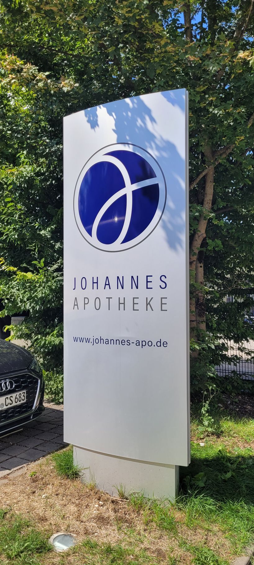 Schild der Johannes Apotheke; blau-weißes Logo auf silbernem Grund, mit grünem Hintergrund aus Bäumen und Gras.