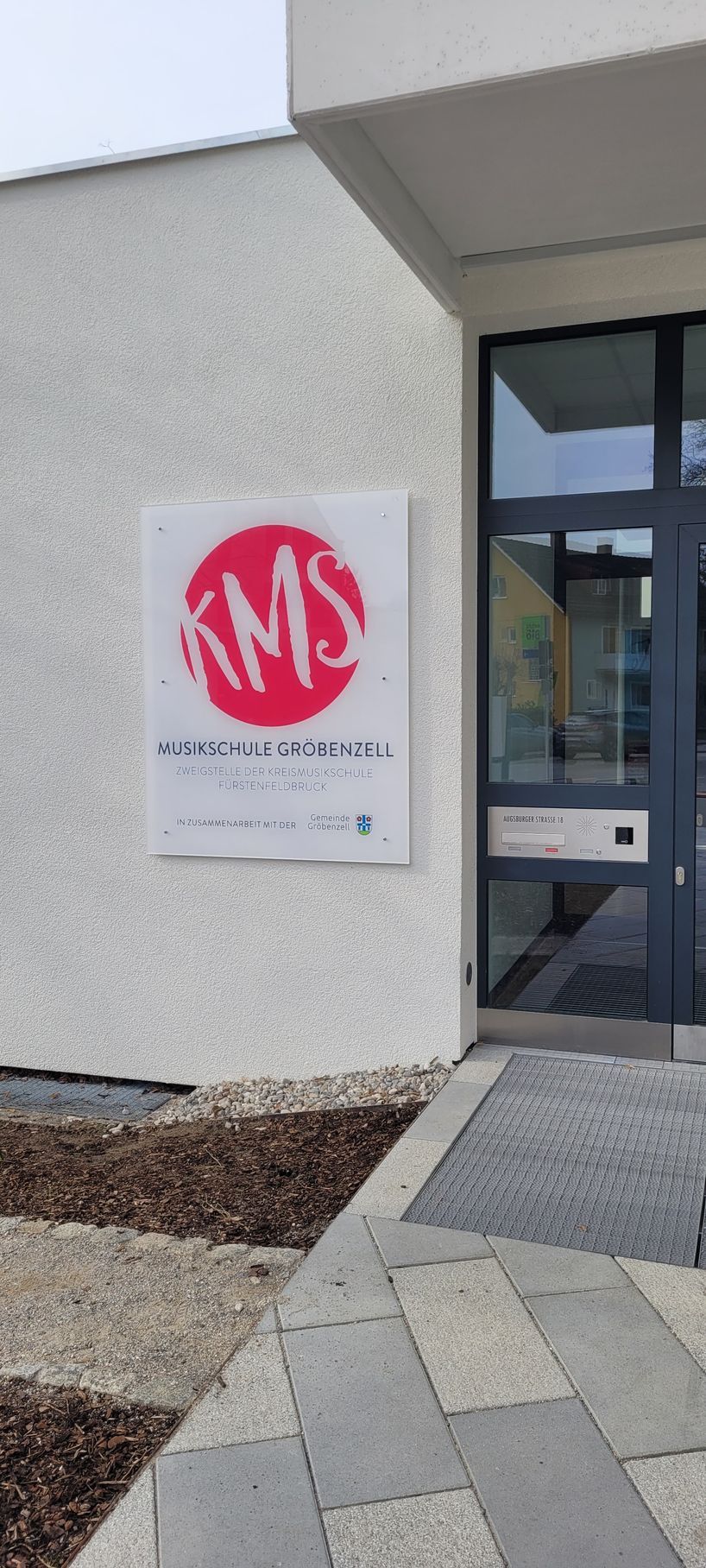 Schild für die KMS-Schule mit einem roten Kreis und weißen Buchstaben an der Außenseite eines Gebäudes mit Steinweg.