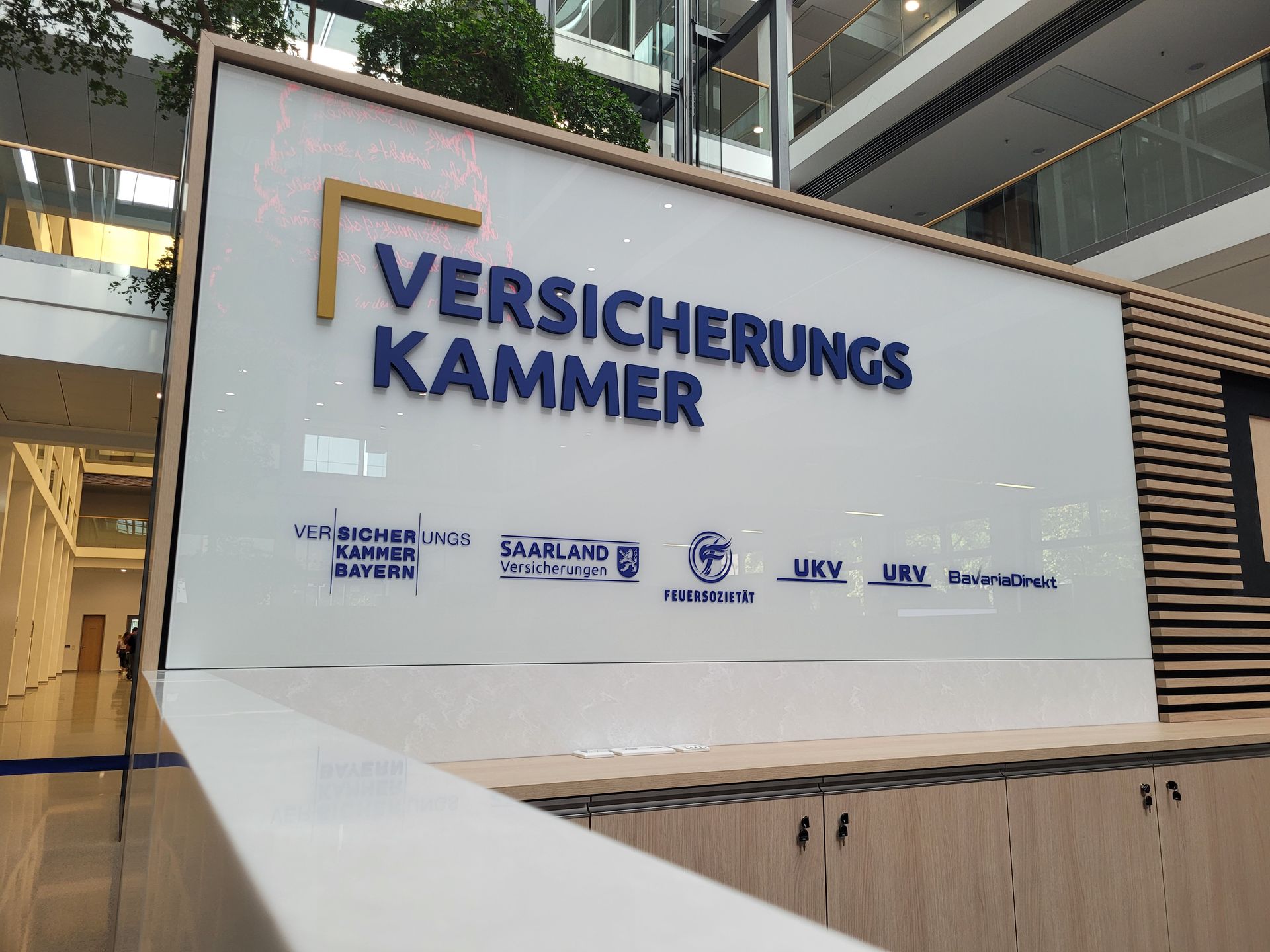 Schild der Versicherungskammer mit blauem Text und Logos in einem modernen Gebäude.