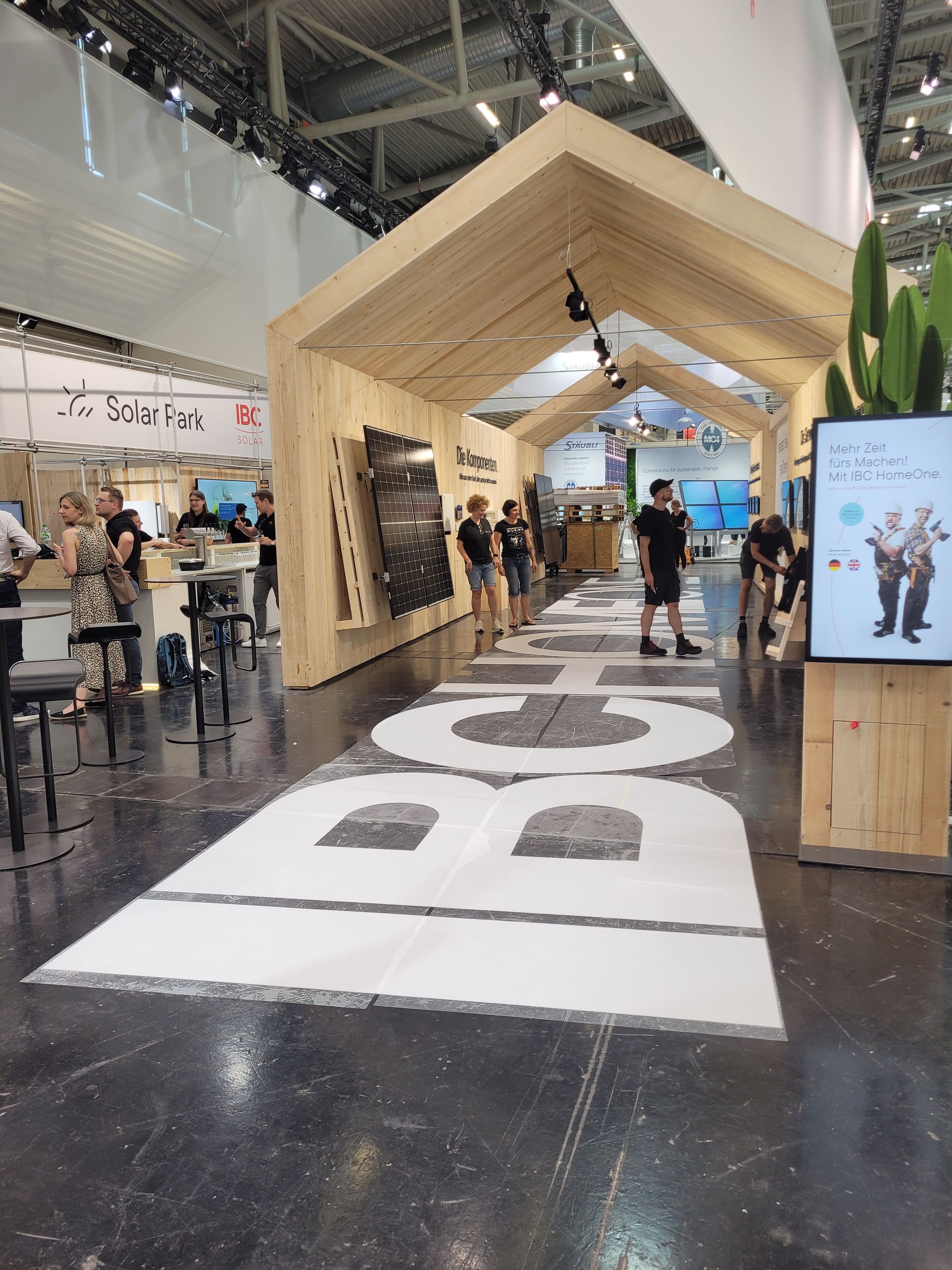 Ein hölzerner Messestand mit großen weißen Buchstaben auf dem Boden. Menschen gehen hindurch.