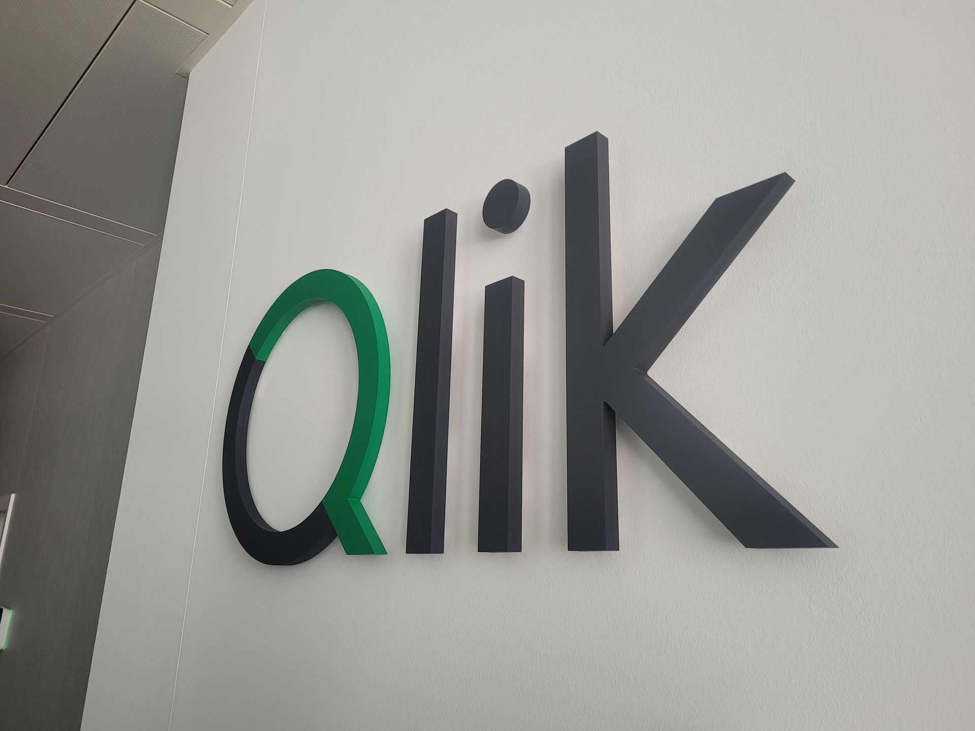 Schwarz-grünes „Qlik“-Logo auf einer weißen Wand; das „Q“ ist grün und schwarz, die anderen Buchstaben sind schwarz.
