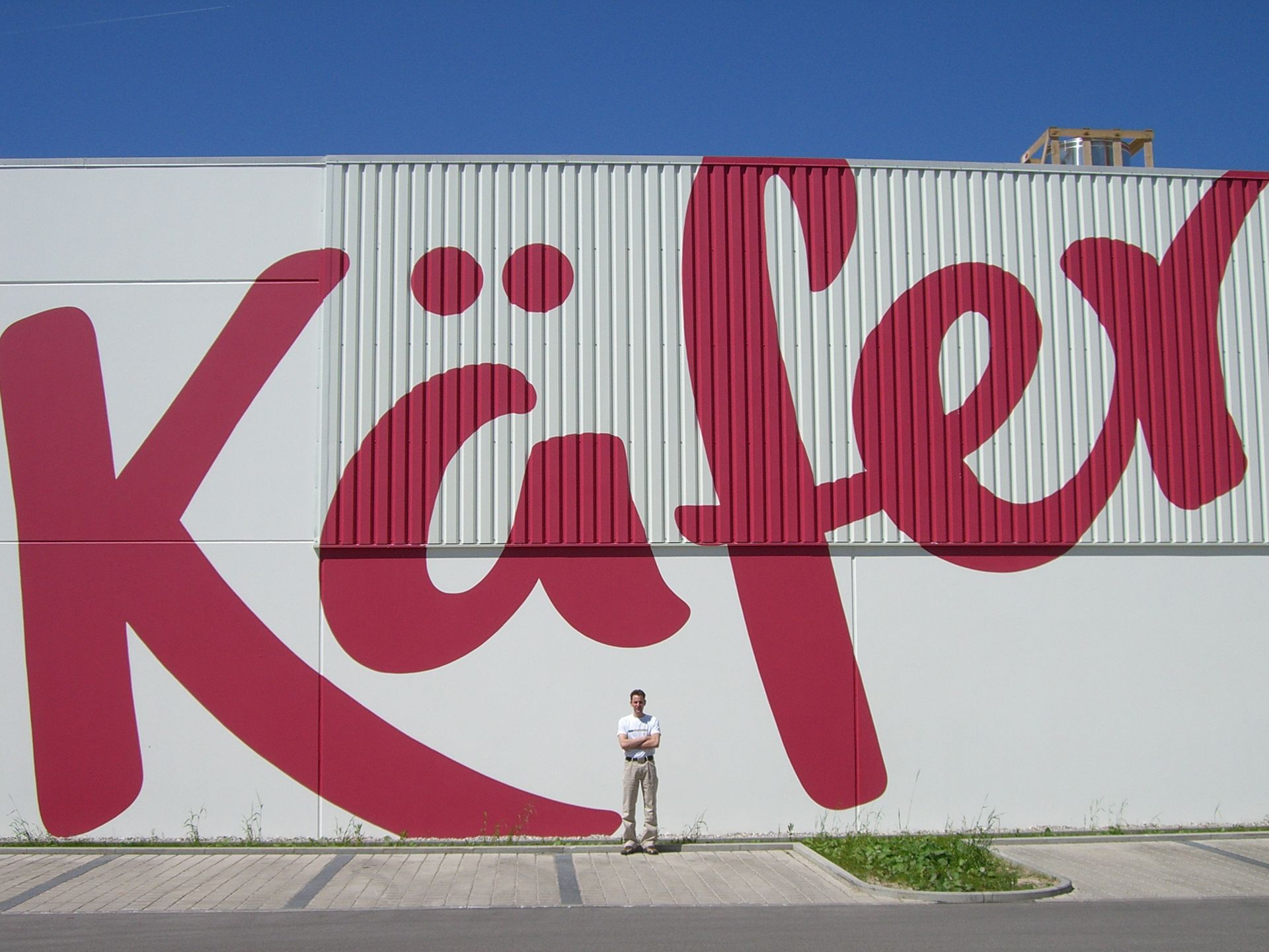 Mann steht vor einem großen roten „Käfer“-Logo auf einer weißen Gebäudefassade, Hintergrund blauer Himmel.