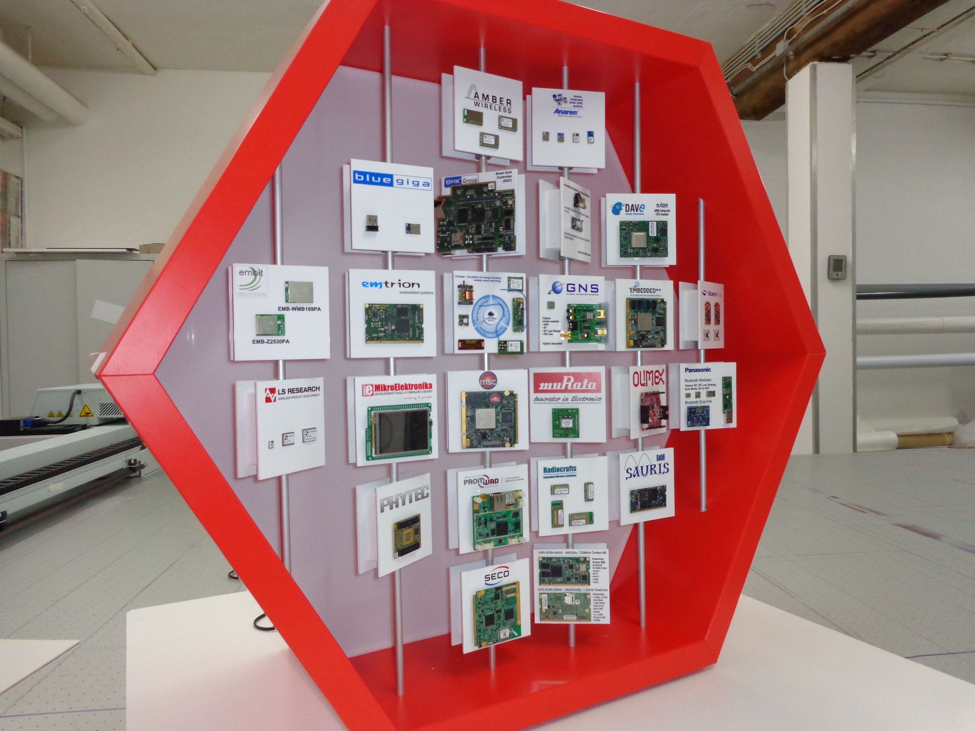 Rote sechseckige Vitrine mit verschiedenen Leiterplatten, mit weißer Rückseite und vertikalen Metallstützen.