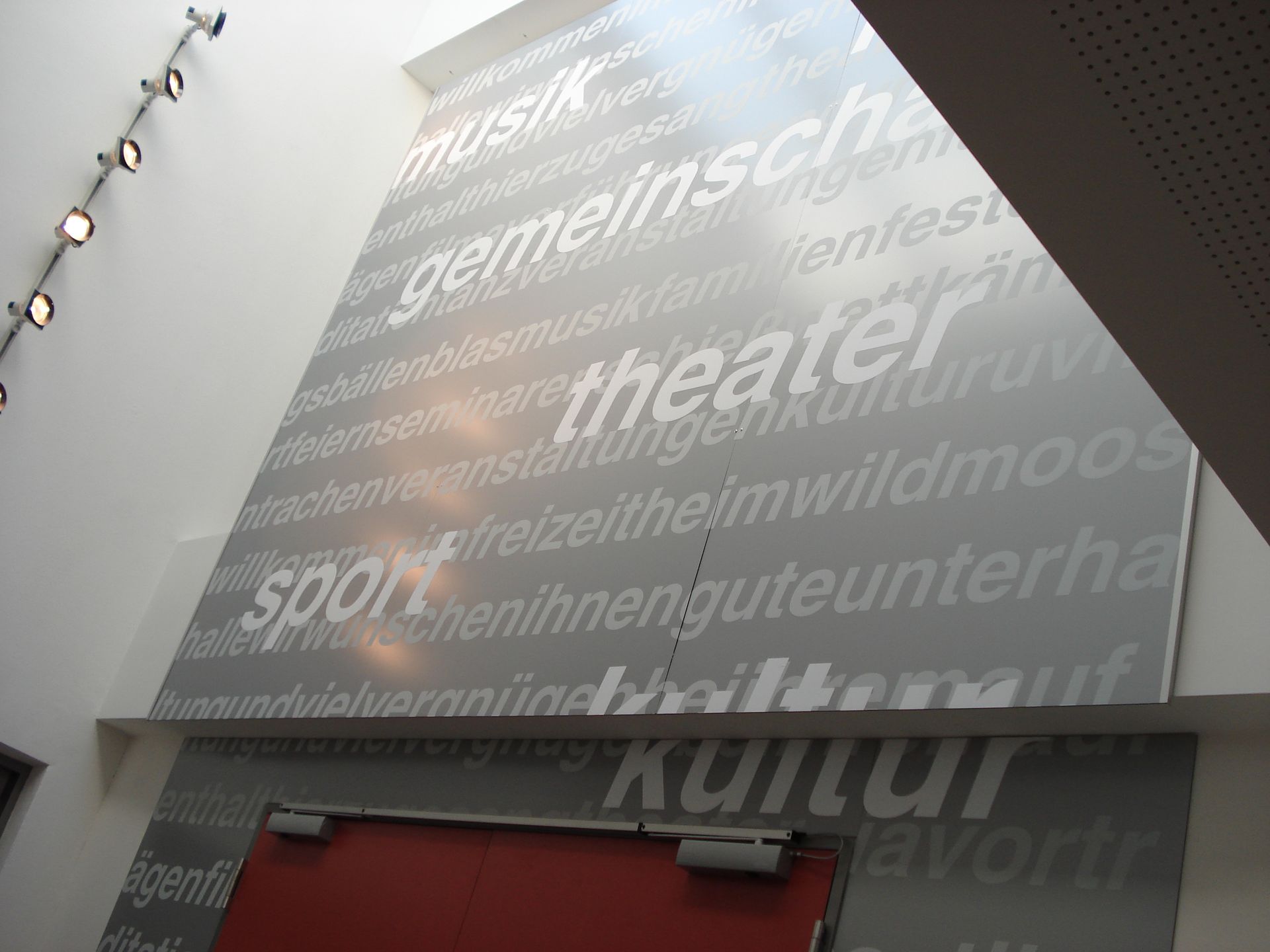 Graue Wand mit Wörtern wie „Musik“ und „Theater“ in weißer Schrift. Darunter ist eine rote Tür.