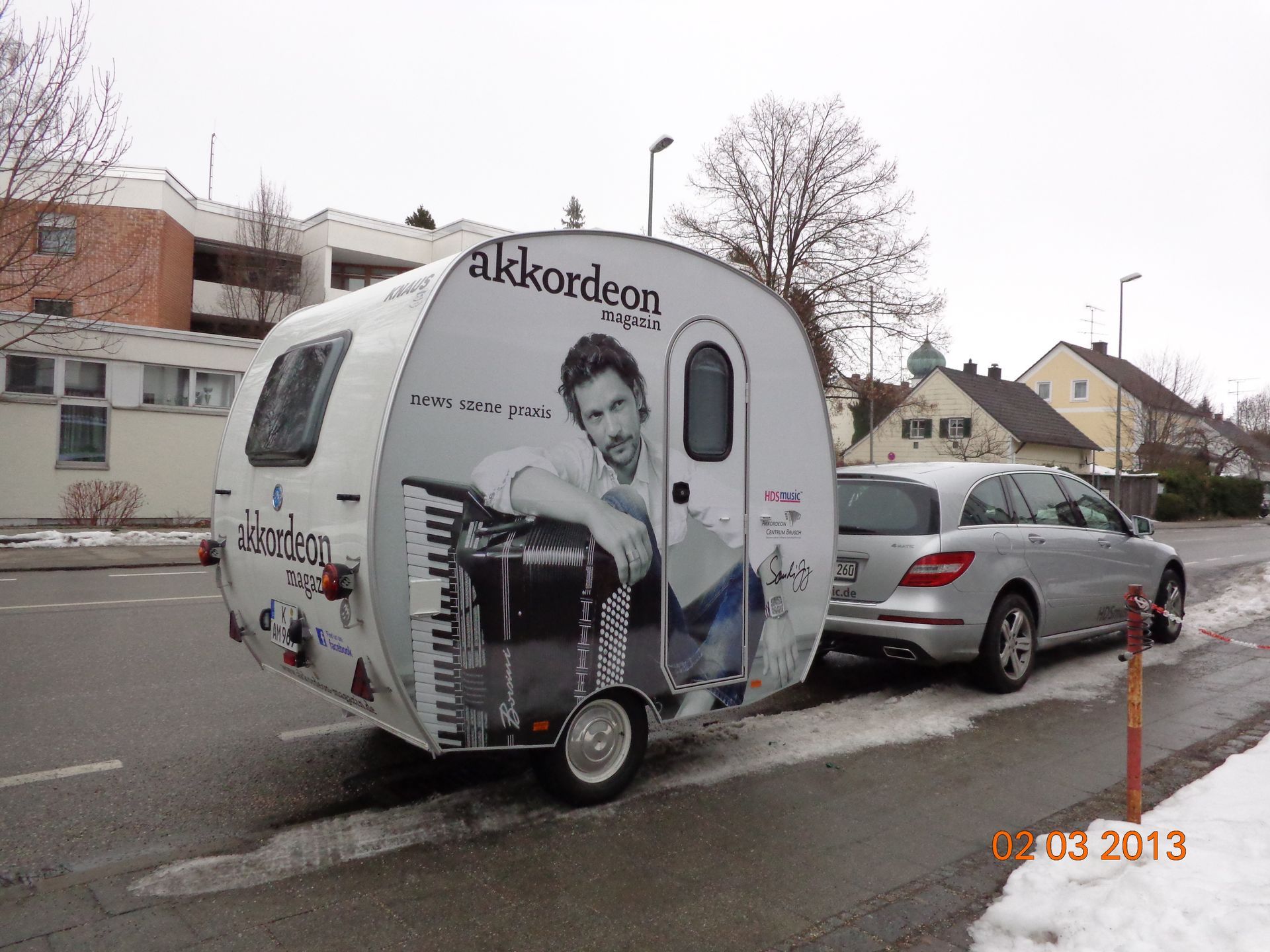 Ein silbernes Auto, das einen weißen Teardrop-Anhänger mit einem Musikerporträt an der Seite zieht.