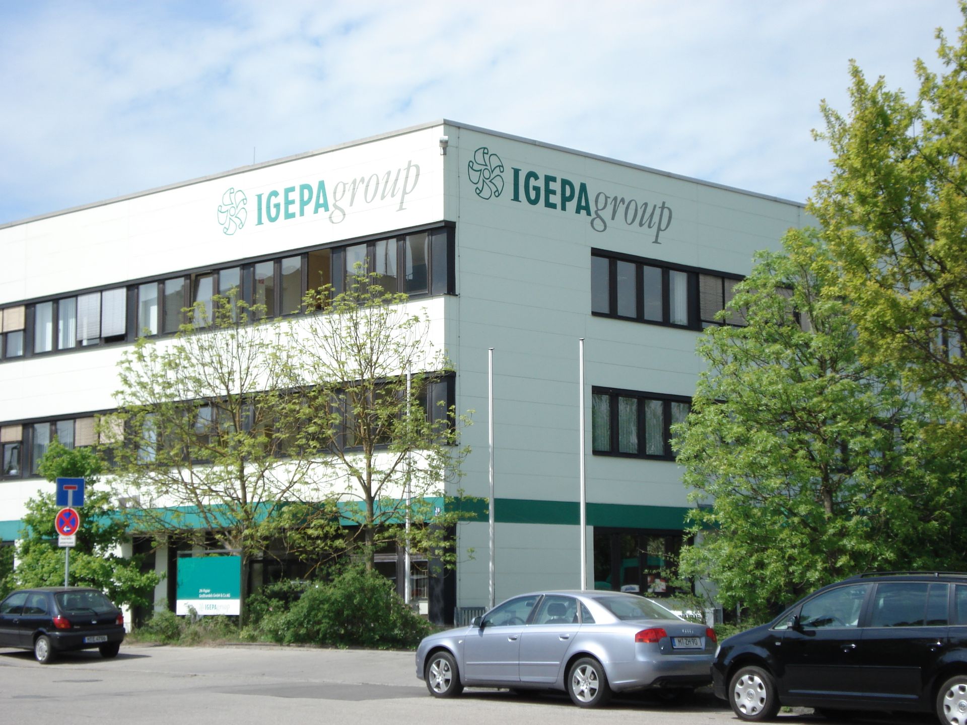 Weißes Bürogebäude mit der Beschilderung „IGEPA Group“. Vor dem Gebäude parken Autos, grüne Bäume.