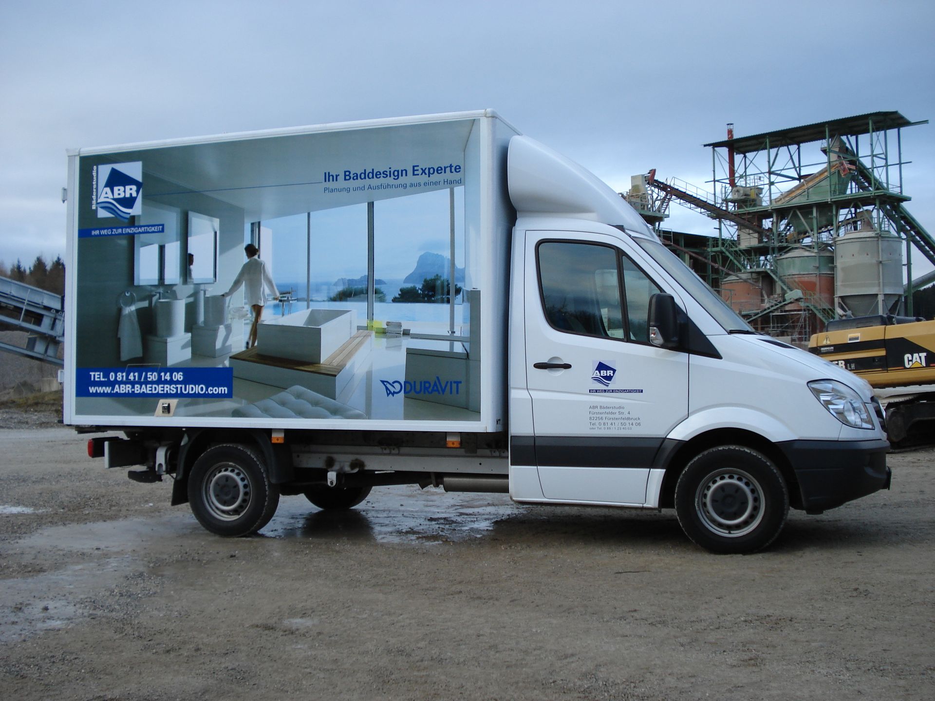 Weißer Lieferwagen mit Werbung für eine Badezimmerszene an der Seite, auf Schotter geparkt.