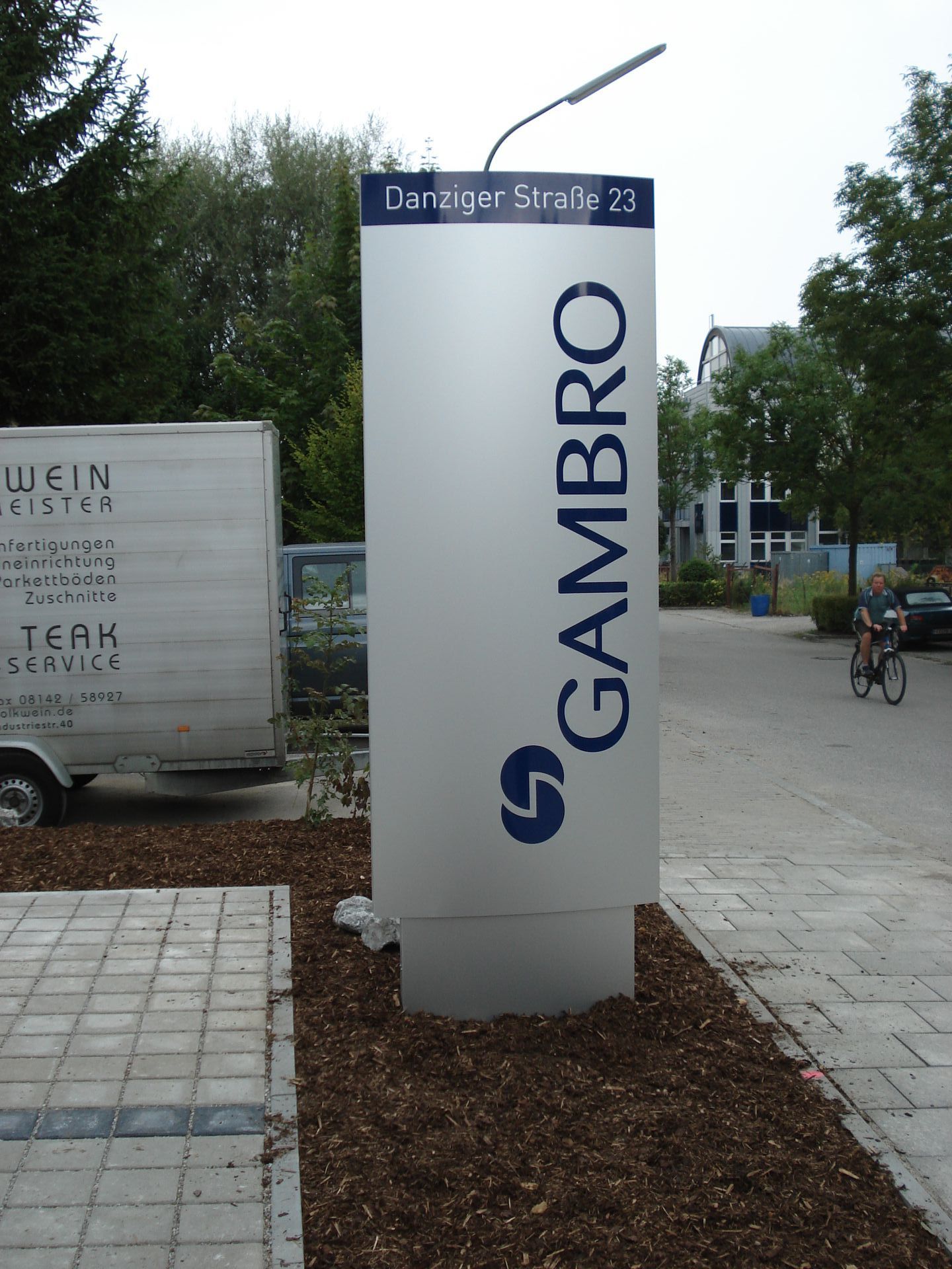 Schild von Gambro in der Danziger Straße 23 mit dem Firmenlogo; im Hintergrund eine Person auf dem Fahrrad.