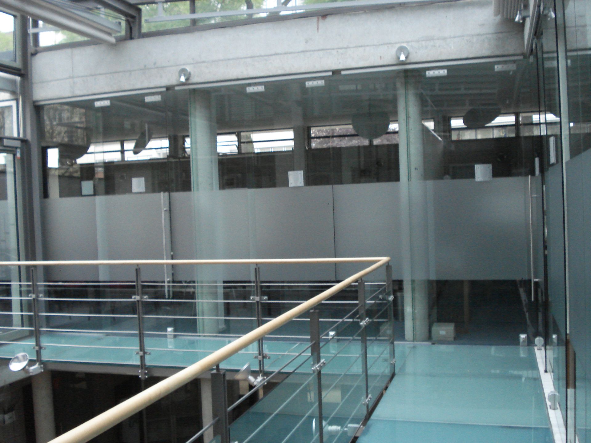 Innenraum mit Glaswänden und einem Gang mit Blick auf eine untere Ebene; Milchglasplatten und Betonakzente.