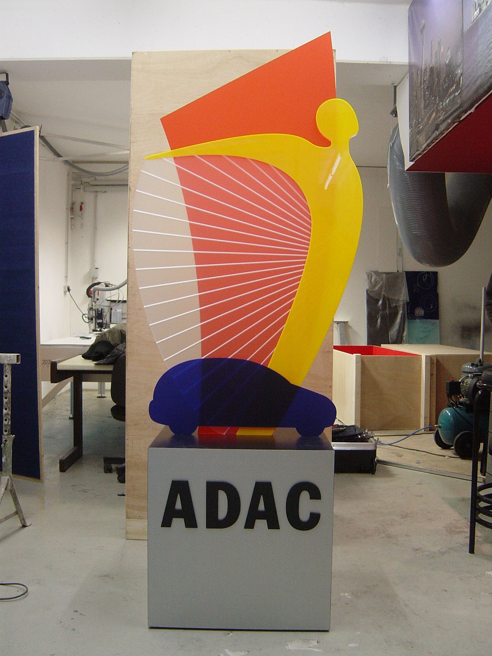 ADAC-Skulptur mit einem blauen Auto auf einem grauen Sockel und einer abstrakten gelb-roten Figur. Werkstatt-Setting.