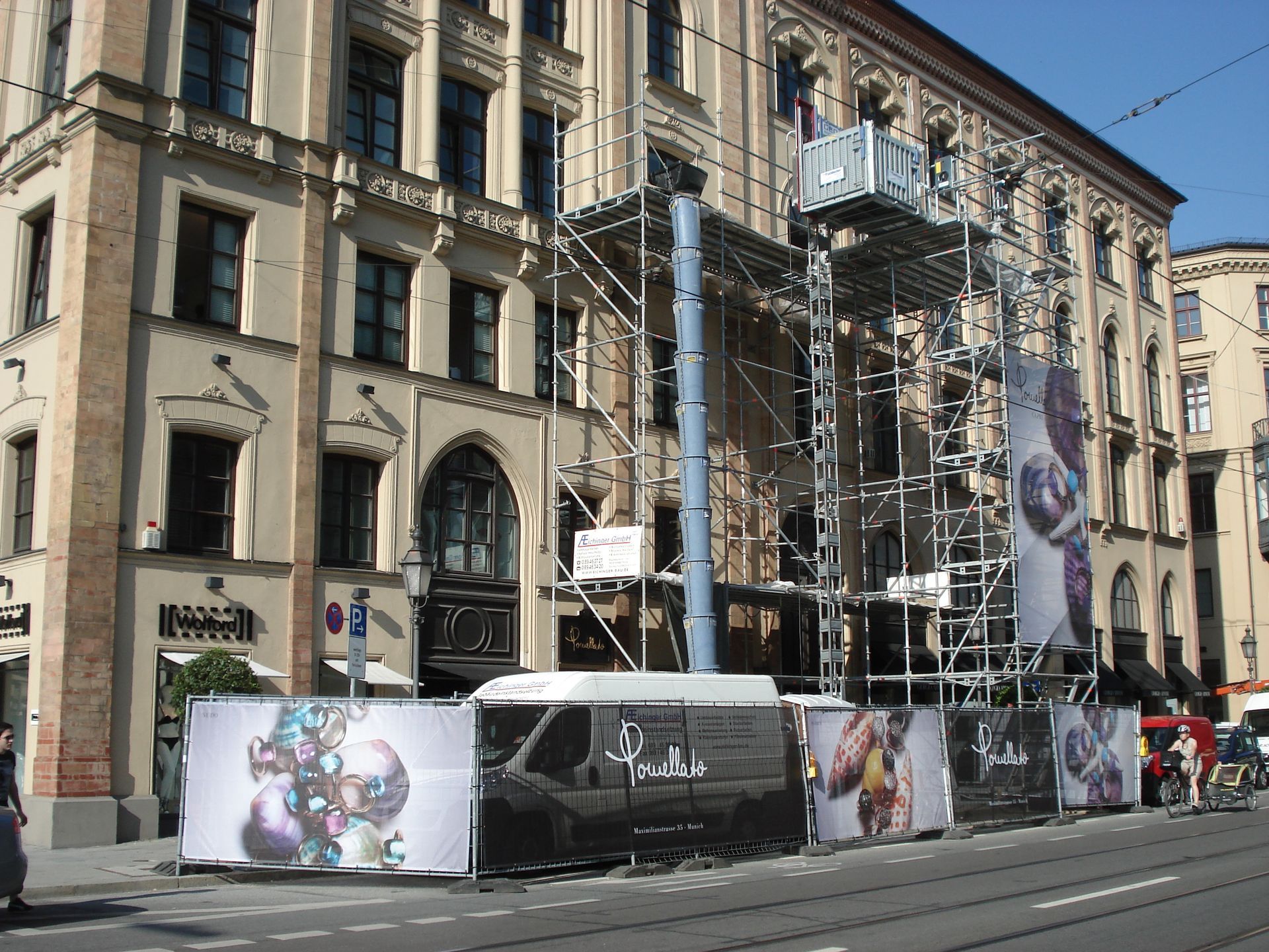 Gerüst vor einem Gebäude mit Bannern, weißem Lieferwagen und Straßenszene.