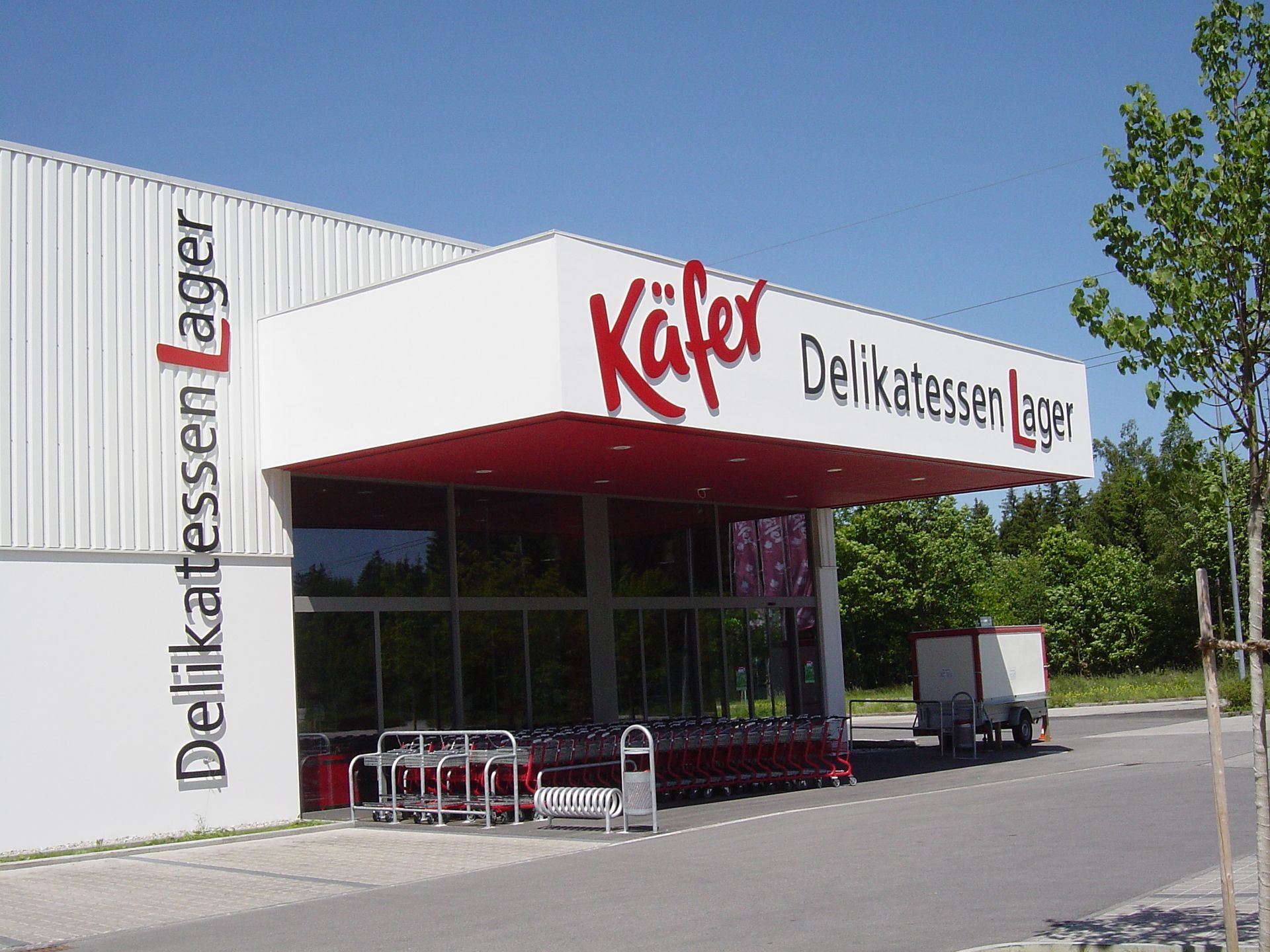 Eingang zum Käfer Delikatessen Lager-Geschäft mit rot-weißer Beschilderung und Einkaufswagen.