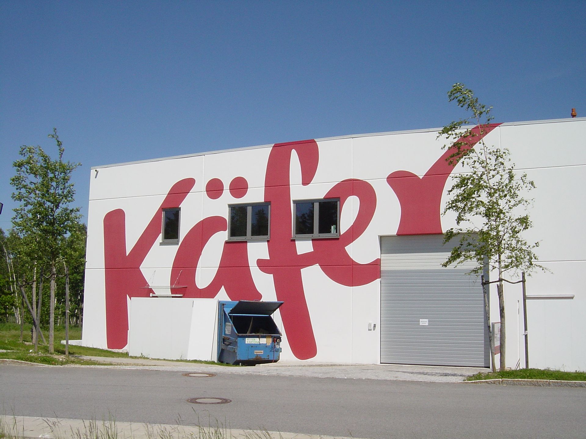 Weißes Gebäude mit großem roten „Käfer“-Logo, darunter parkendes blaues Lieferfahrzeug, blauer Himmel.