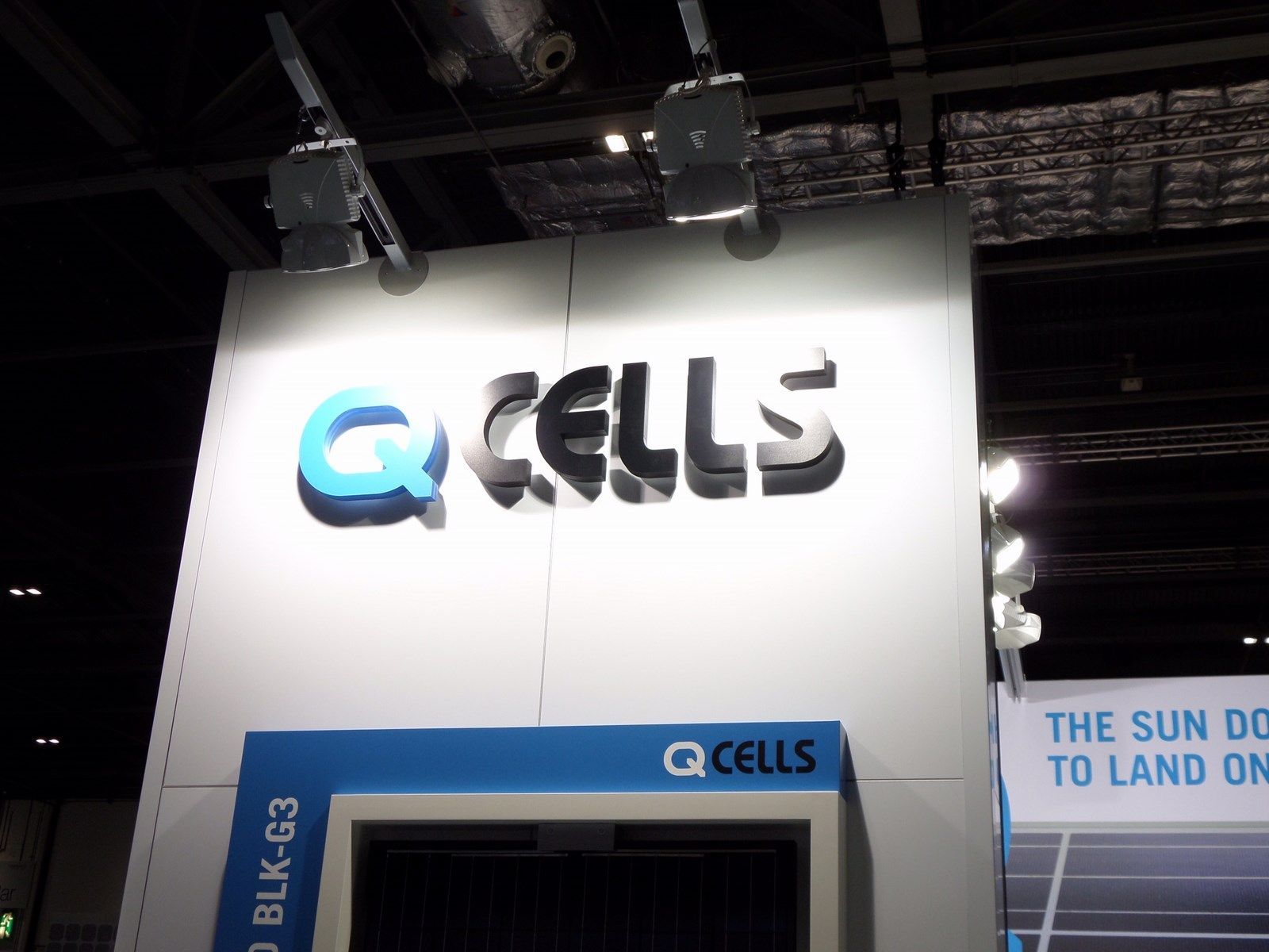 Q CELLS-Logo auf einer weißen Wand mit blauen Akzenten und Scheinwerfern. Unten ist eine Produktanzeige zu sehen.