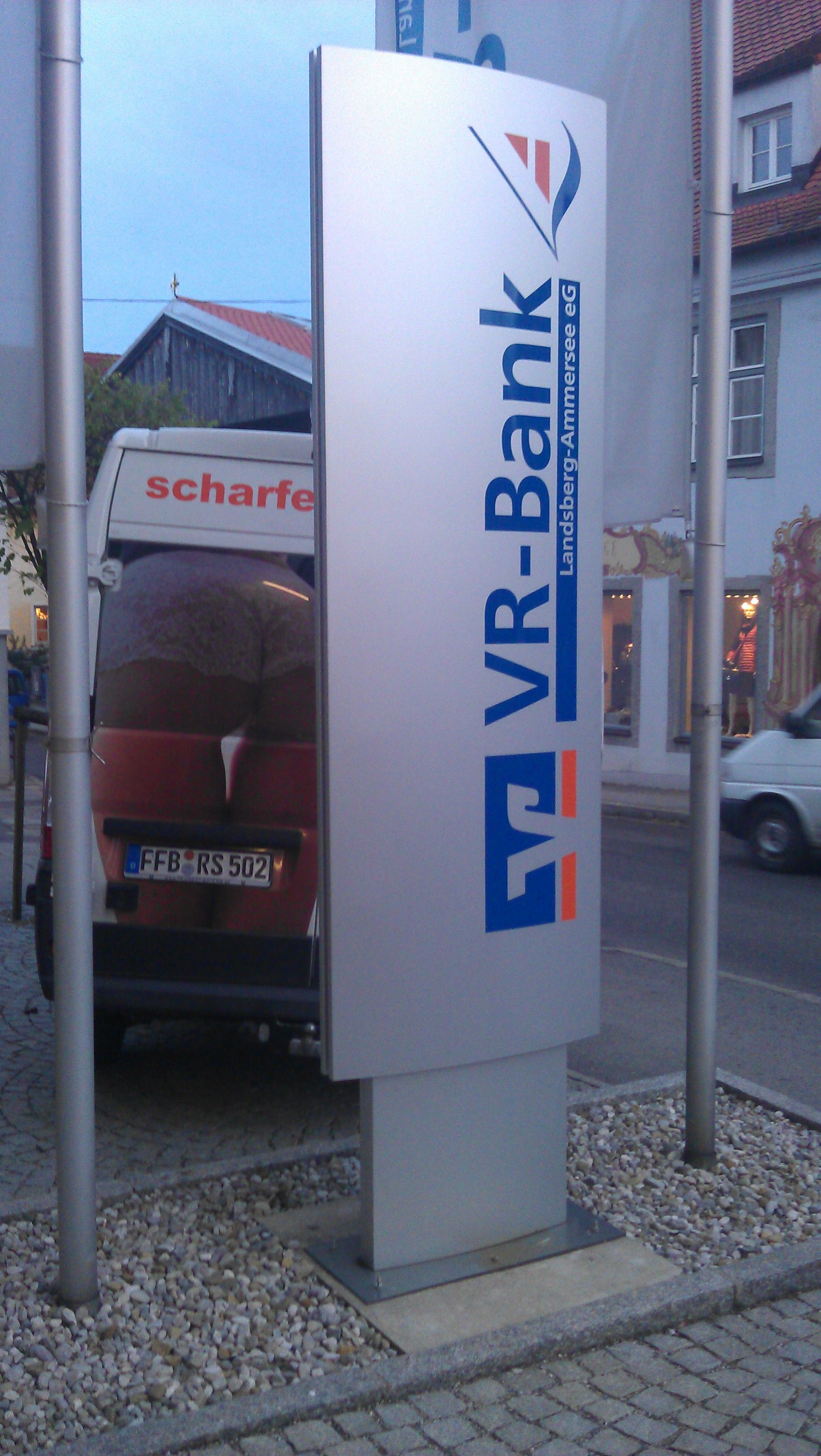 VR-Bank-Schild, blau-oranges Logo auf silbernem Pylon, im Außenbereich.