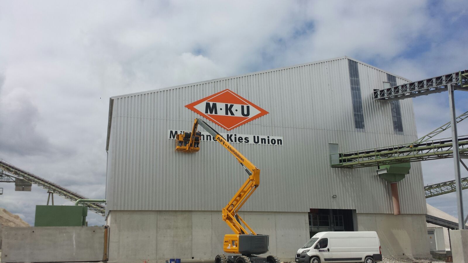 Ein Arbeiter auf einer Hubarbeitsbühne malt das Logo „MKU Mineração Unidos“ auf ein großes Industriegebäude.