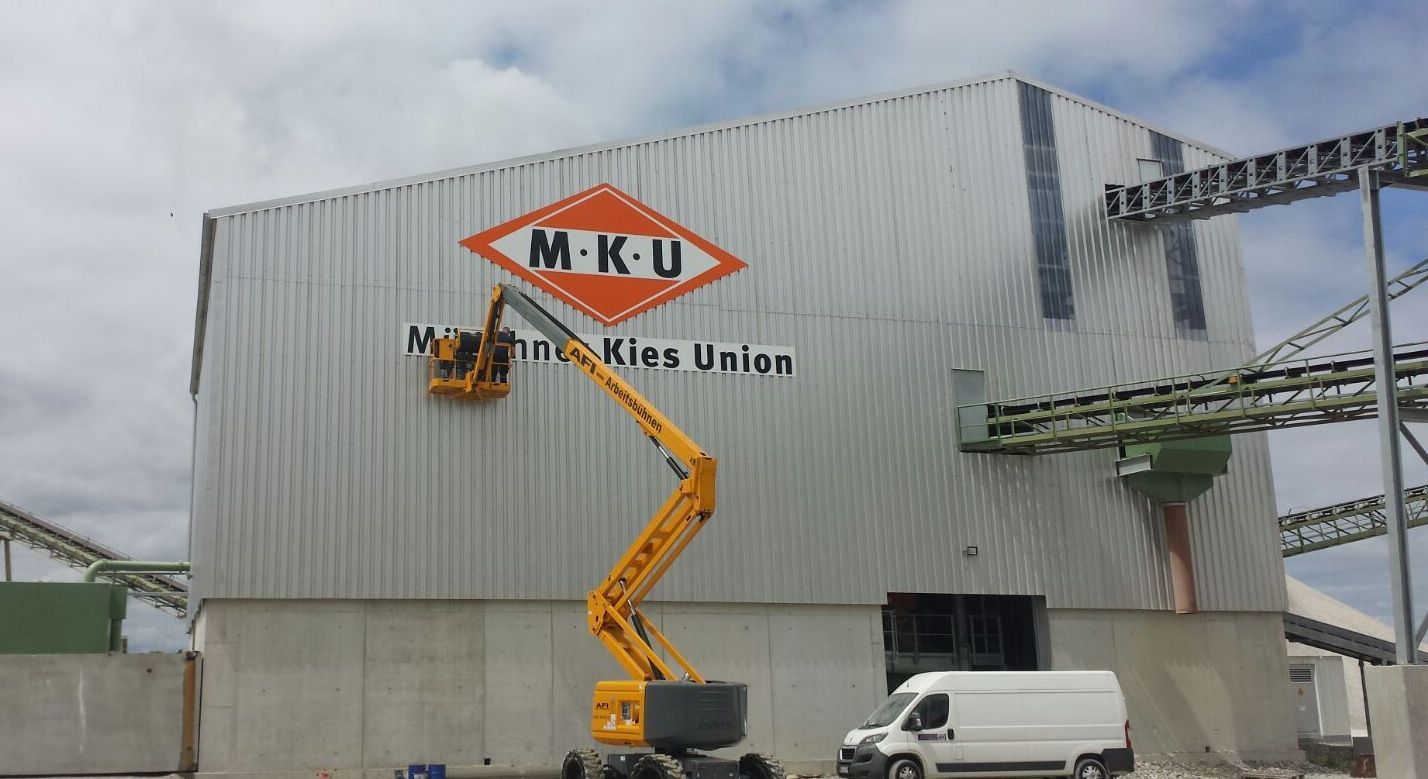 Arbeiter im Kran befestigen das MKU-Logo auf die Außenseite eines Industriegebäudes.