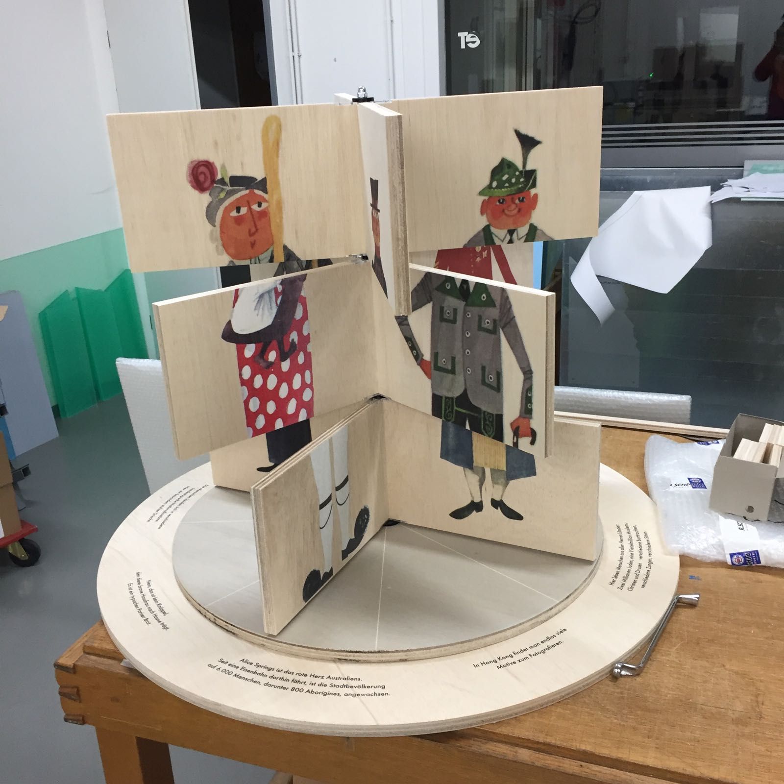 Holzskulptur mit abgebildeten Figuren in traditioneller Kleidung. Auf einer Drehplattform in einer Werkstatt aufgestellt.