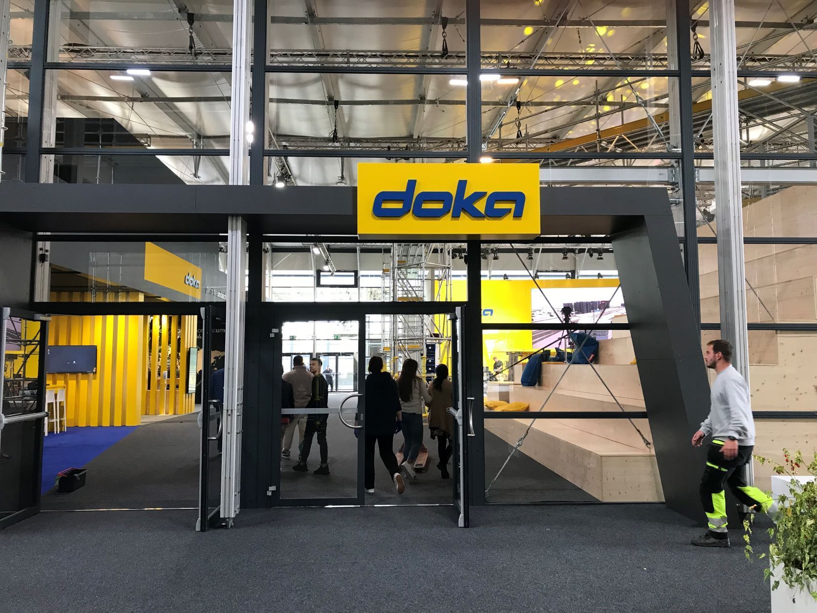 Eingang zu einem „Doka“-Stand auf einer Messe; Menschen betreten/verlassen den Stand. Gelbes Logo, graue und gelbe Akzente.