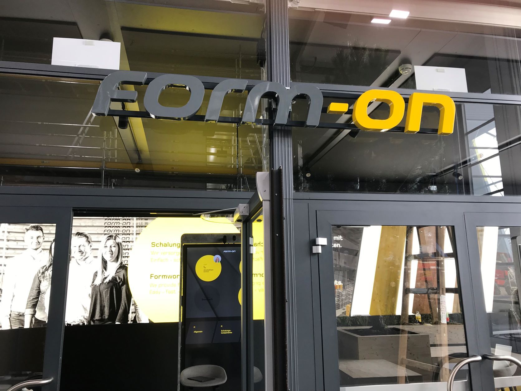 Eingang zum „form-on“-Laden; grau-gelbes Schild über Glastüren; im Inneren ist ein Schwarz-Weiß-Bild zu sehen.