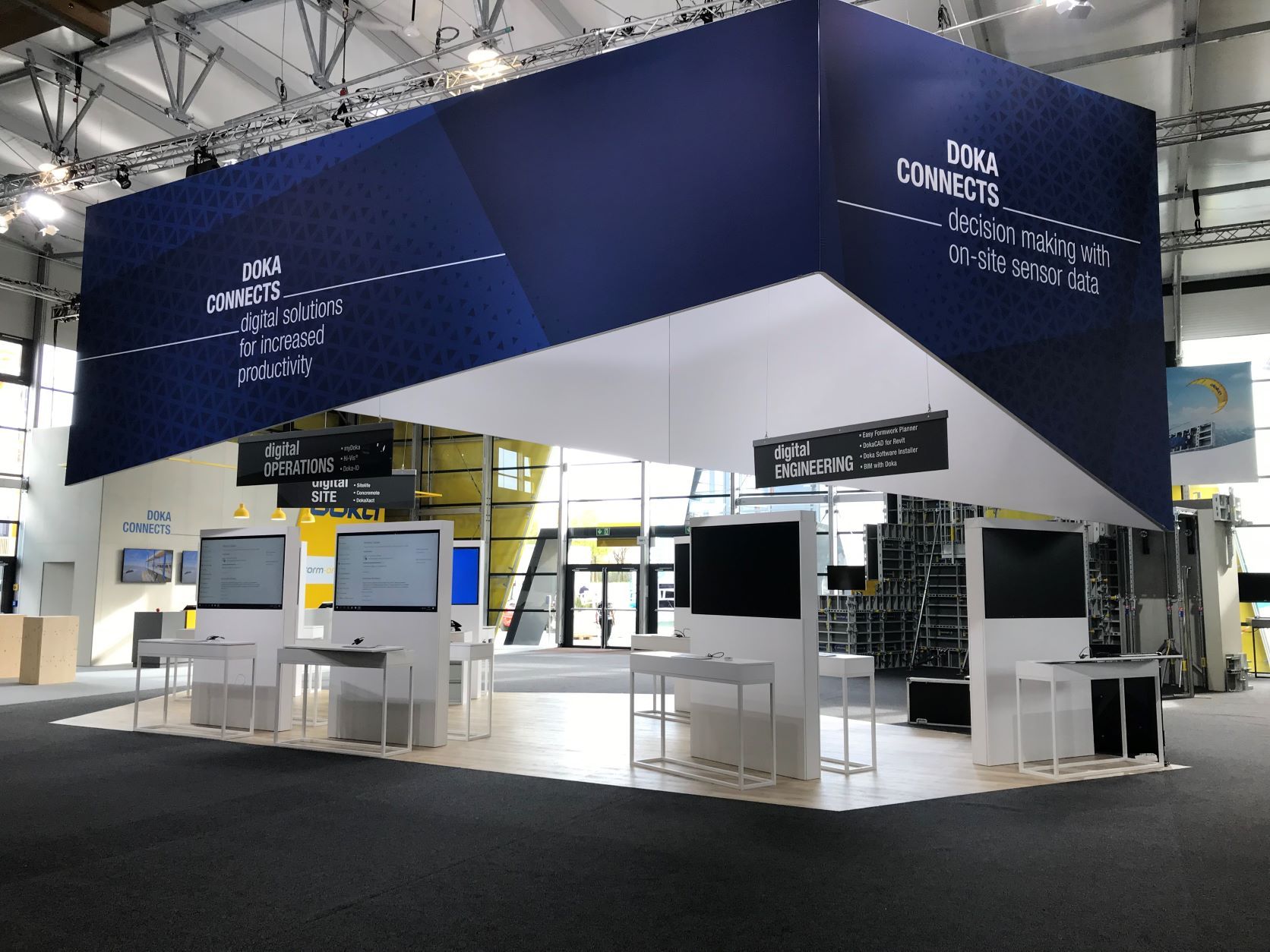 Messestand mit dunkelblauem Banner und weißen Kiosken. Der Text auf dem Banner lautet „Doka Connects“.