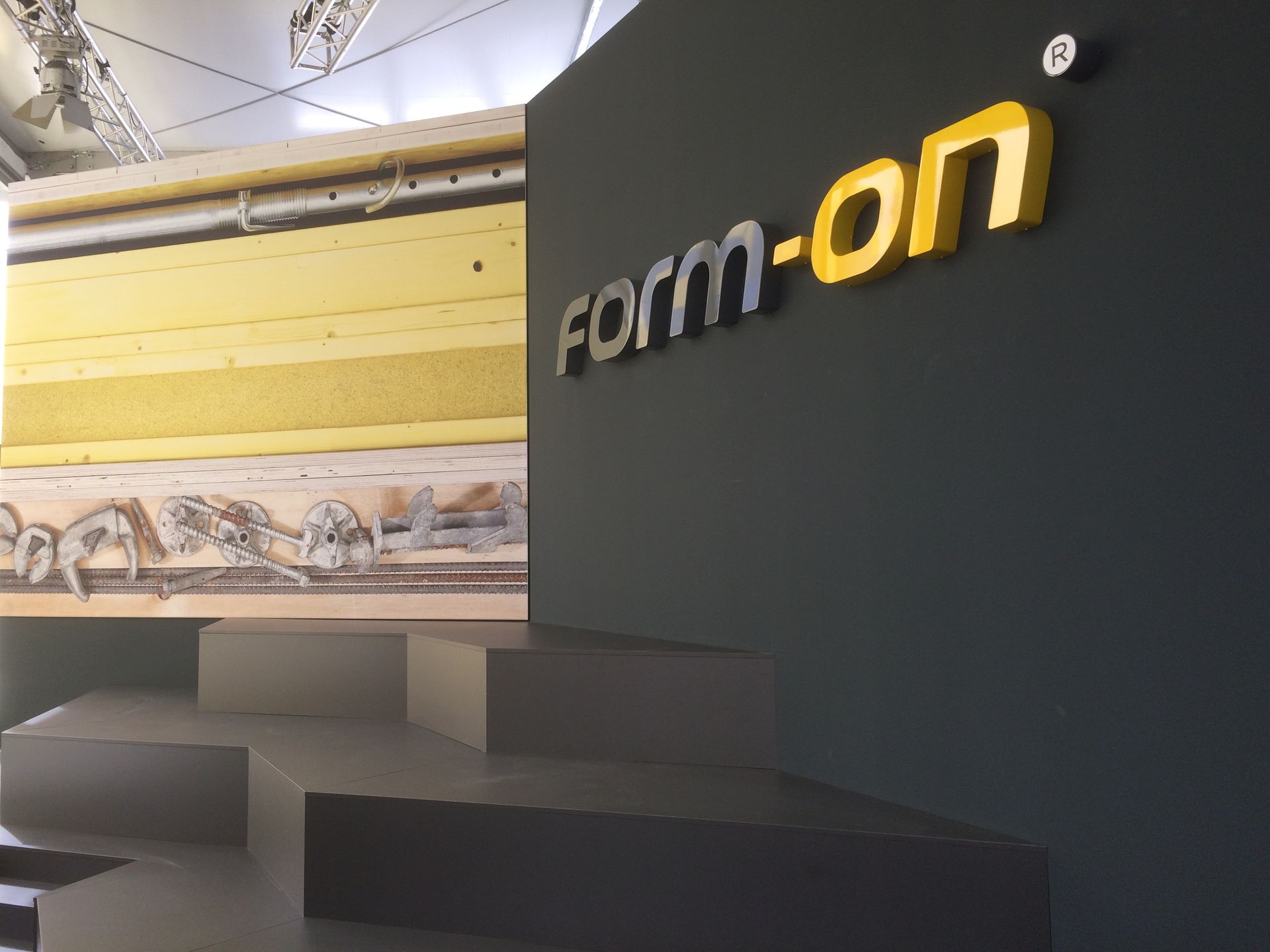 Dunkelgraue Wand mit gelbem „Form-on“-Schild und grauen Stufen, im Hintergrund eine geschichtete Materialpräsentation.