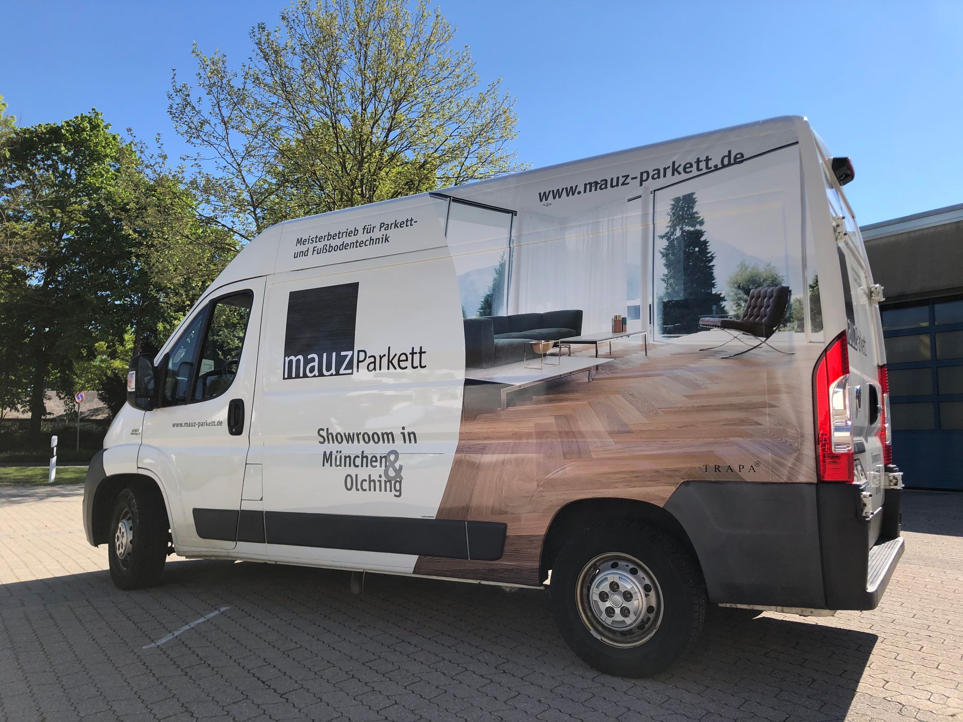 Weißer Lieferwagen mit Werbung auf Holzboden, vor einem Gebäude geparkt.
