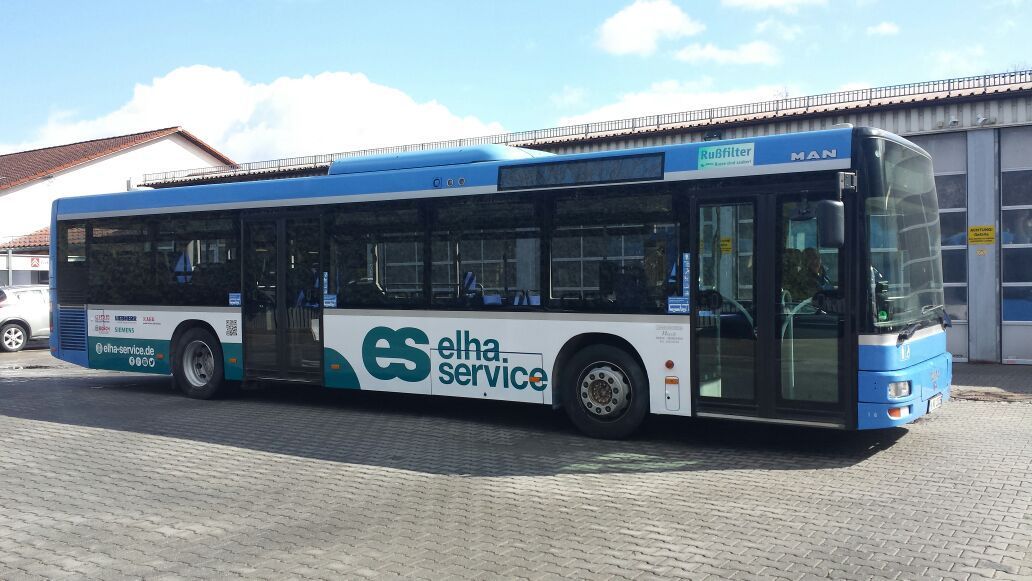 Ein blau-weißer Bus des „Elha-Dienstes“ parkt an einem sonnigen Tag vor einem Gebäude.