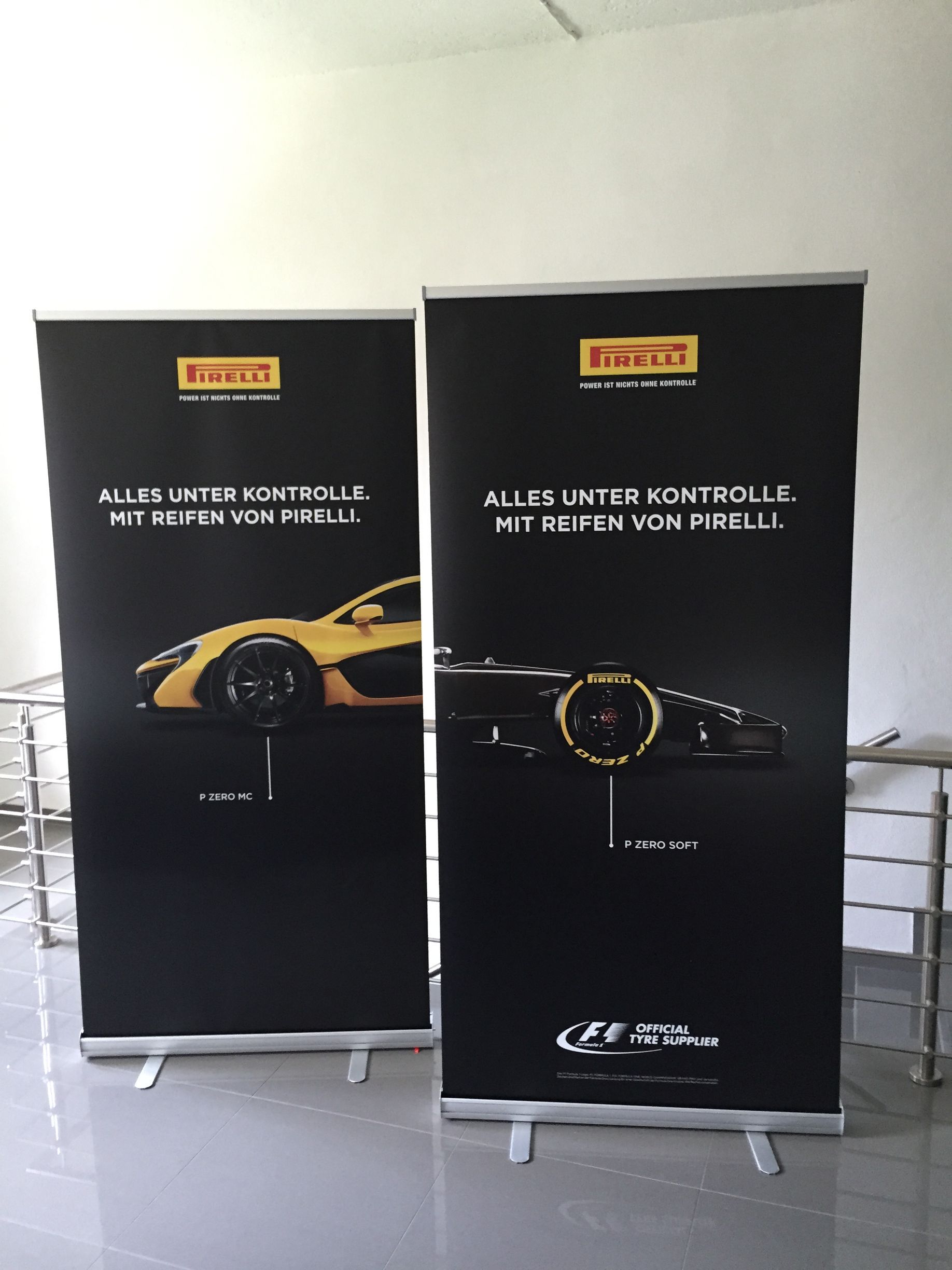 Zwei schwarze Pirelli-Banner mit goldenen Logos, die ein goldenes und ein graues Auto zeigen.