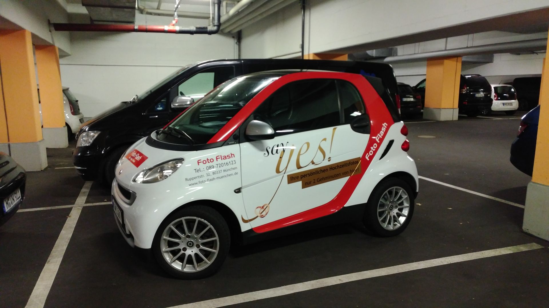 Smart-Auto mit Werbung in einem Parkhaus geparkt. Weiß-rotes Farbschema.
