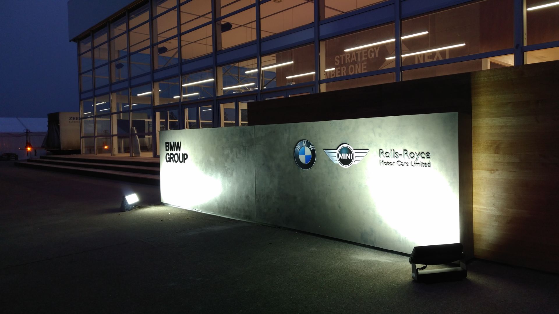 Eingang zum Gebäude der BMW Group bei Nacht, beleuchtetes Schild mit den Logos von BMW und Rolls-Royce.