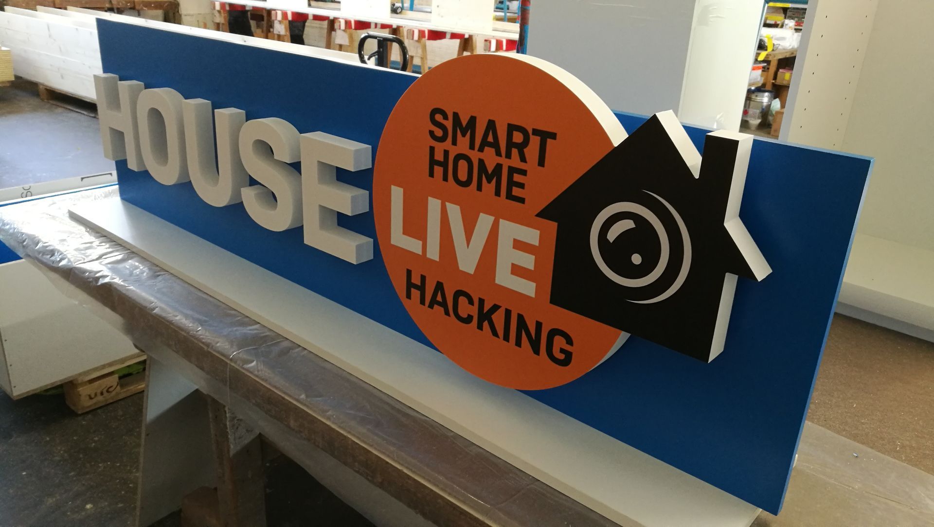 Schild für „Smart Home Live Hacking“ auf blauem Hintergrund mit orangefarbenem Kreis und Hausgrafik.