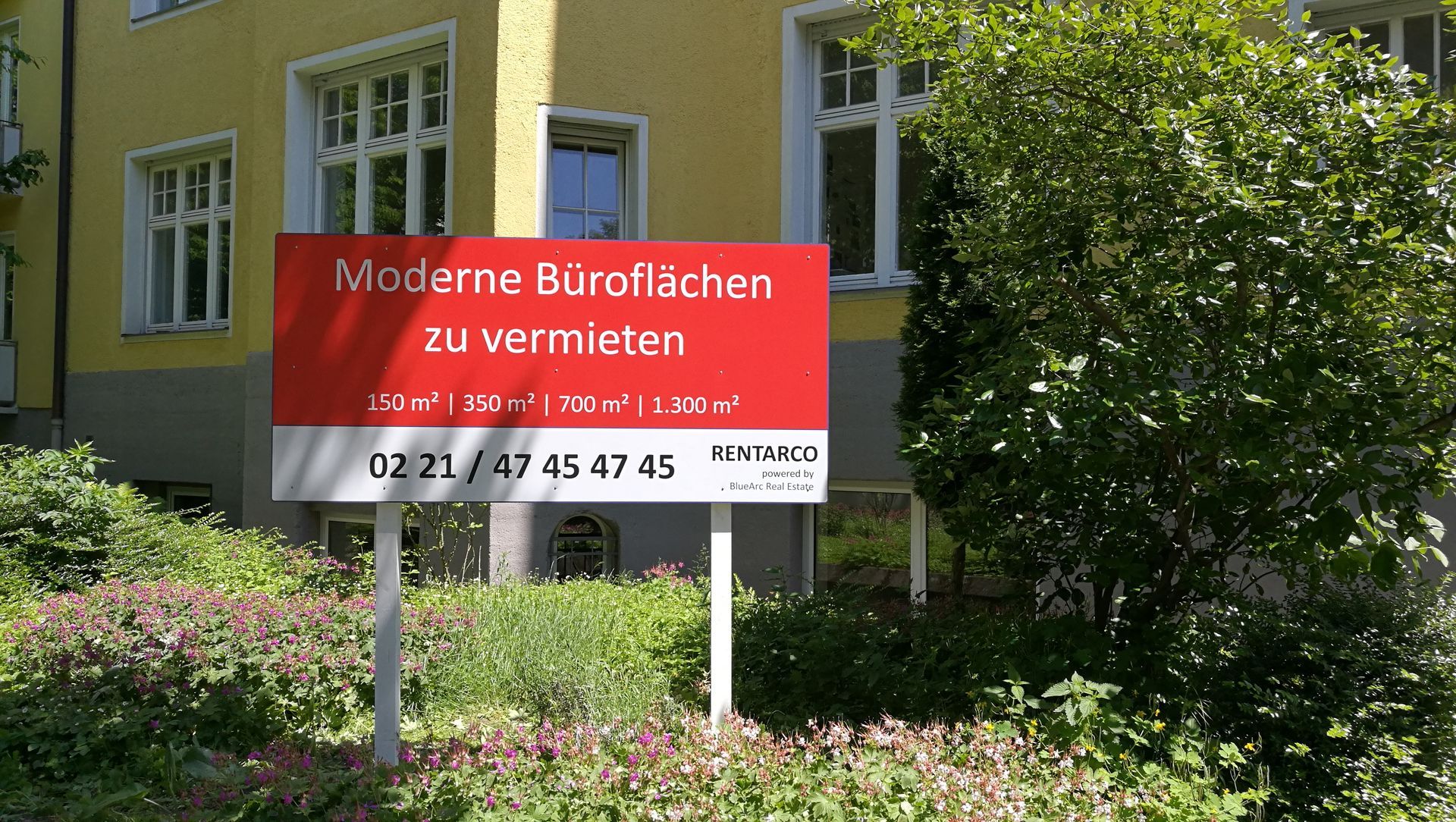 Rotes Schild mit der Aufschrift „Moderne Büroräume zu vermieten“ vor einem gelben Gebäude, grüne Büsche.